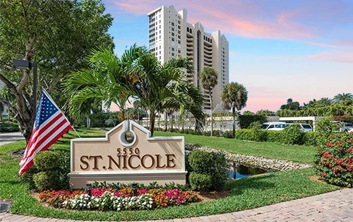 Sign for St. Nicole with American flag, surrounded by flowers and palm trees, in front of tall buildings, during sunset.
