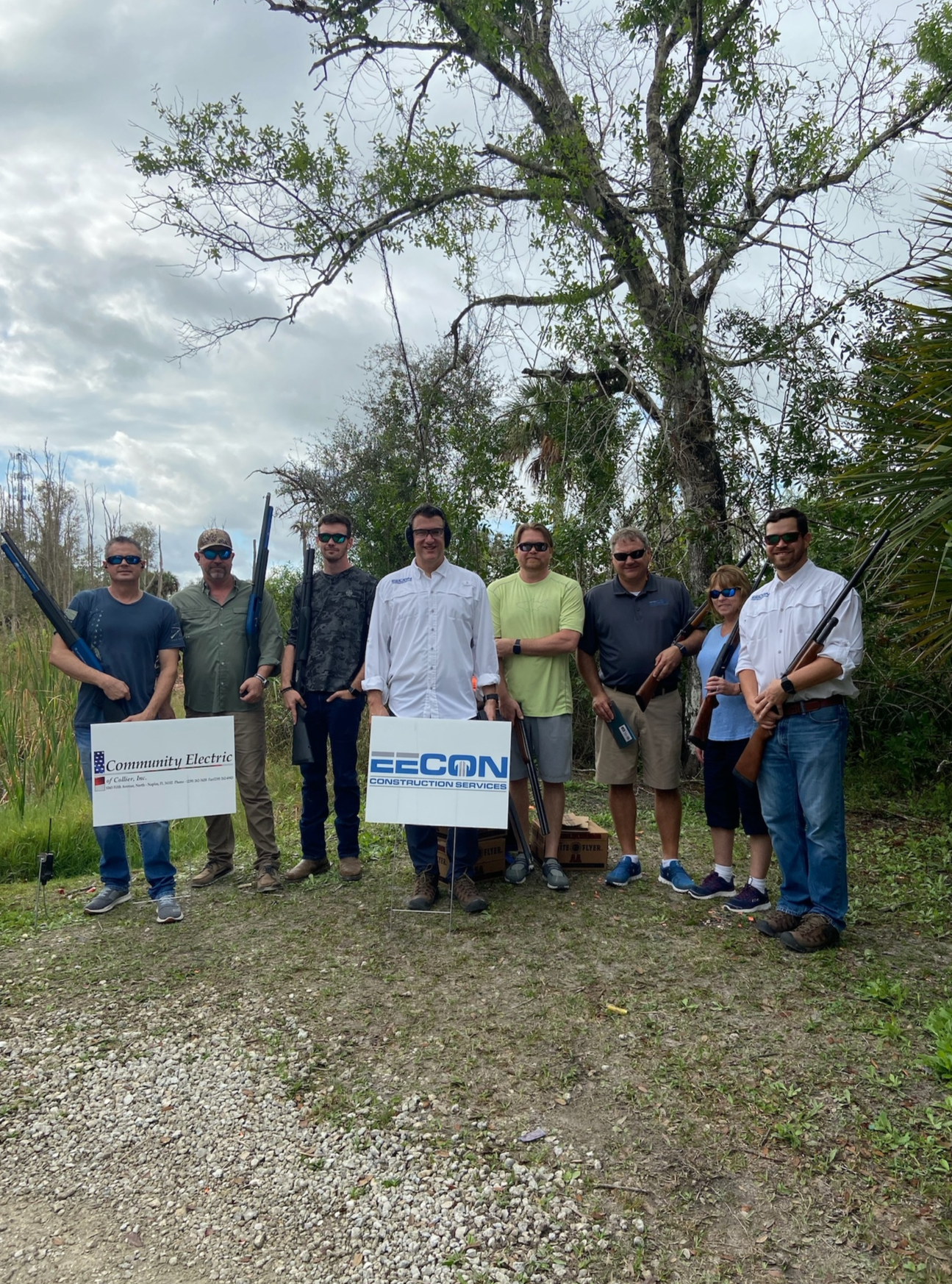 Group of eight people outdoors holding shotguns, standing behind signs for Community Electric and EECON Construction Services, with trees and cloudy sky in the background.