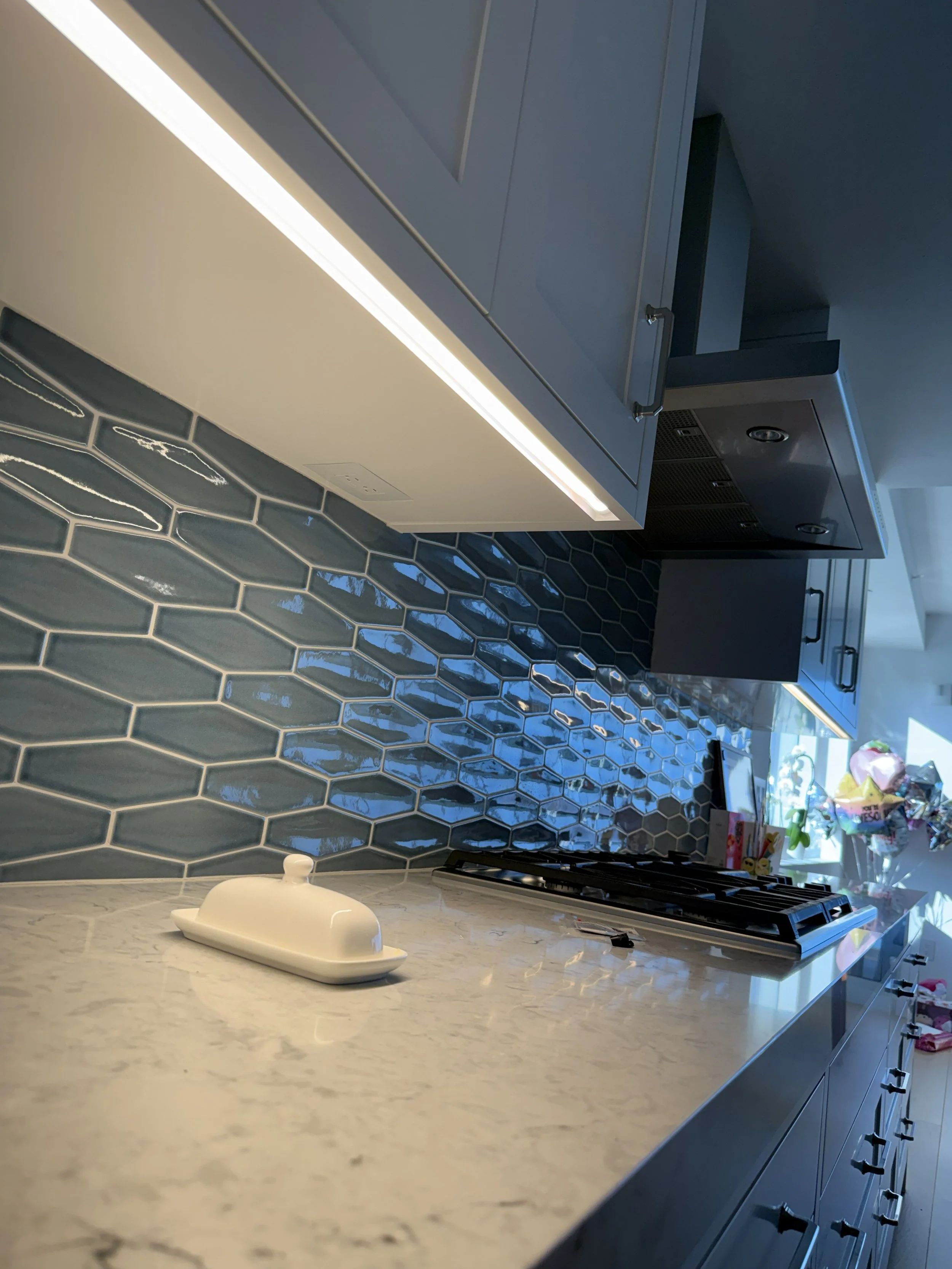 A modern kitchen with a marble countertop, navy blue tile backsplash, and gray cabinets. There is a white tray on the counter and a gas stove below the cabinets.