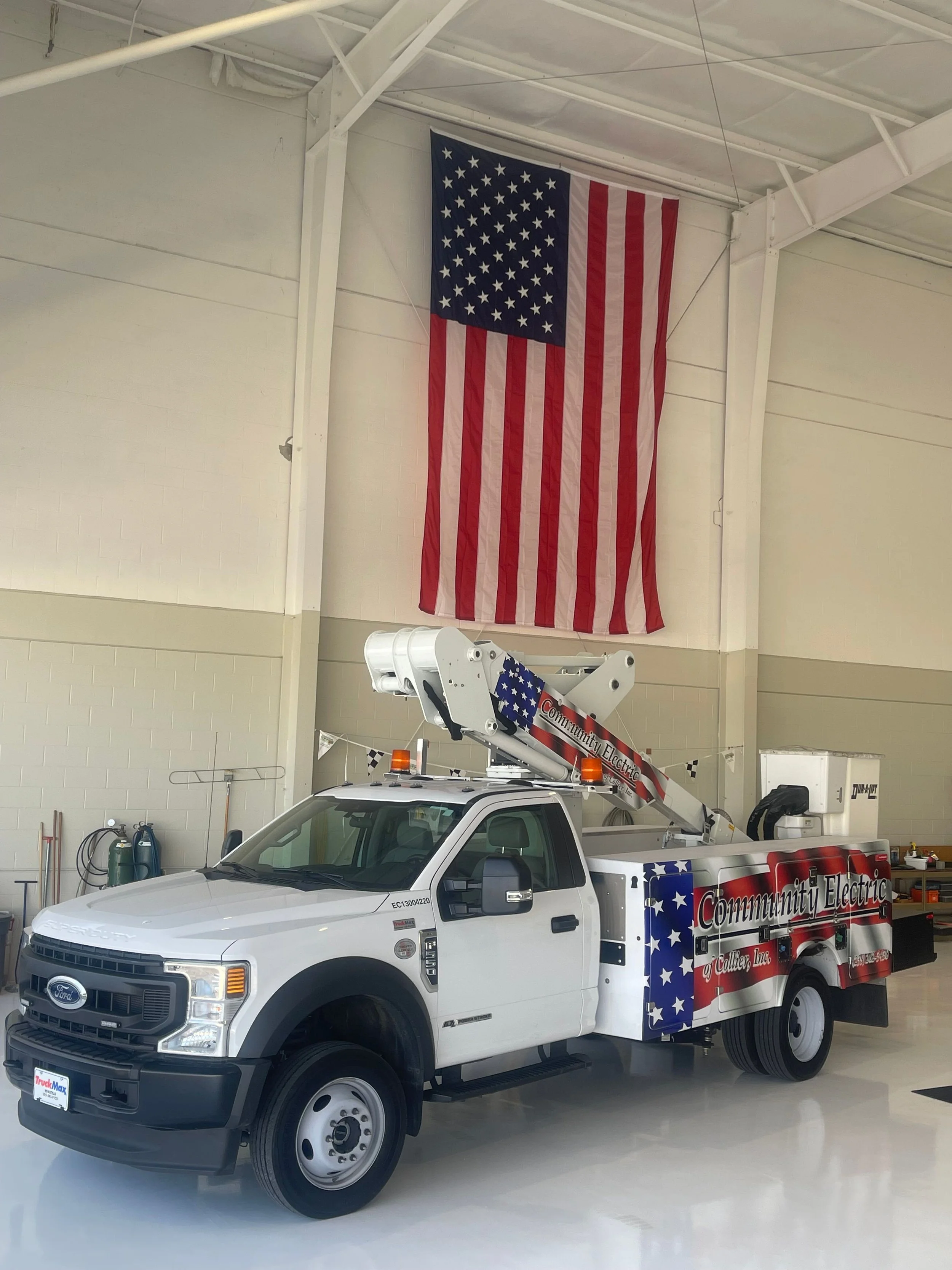 A white utility truck with American flag graphics and the text 'Community Electric' on the side. The truck is inside a large, well-lit industrial building with a high ceiling. A large American flag hangs from the ceiling in the background.