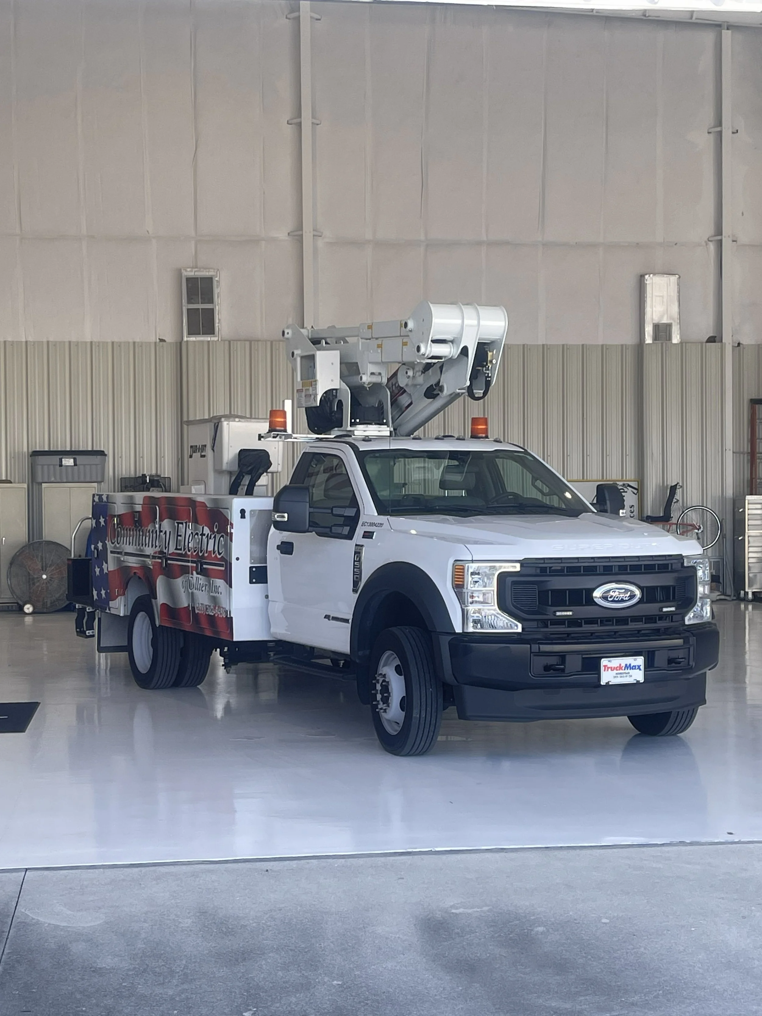 A white utility truck with a bucket lift, parked indoors on a shiny floor. The truck has the logo 'Community Electric Jollier LLC' on the side and is equipped with orange warning lights on top.