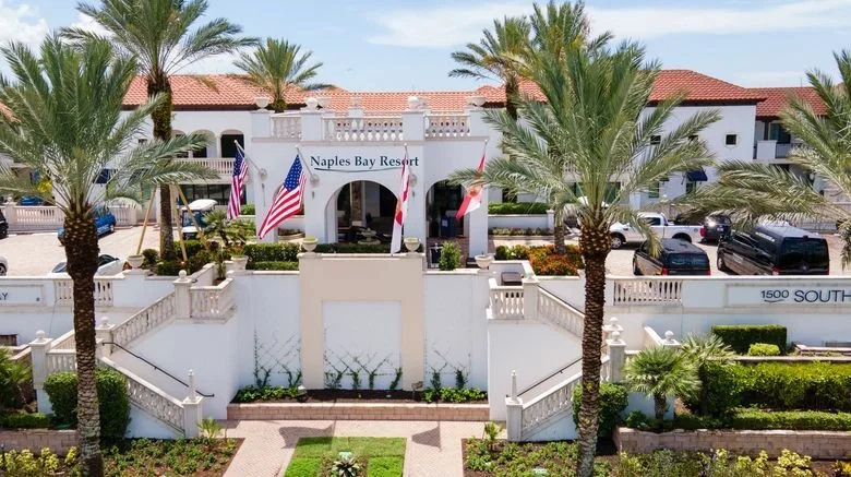 Exterior view of Naples Bay Resort with palm trees, flags, and parking lot in front.