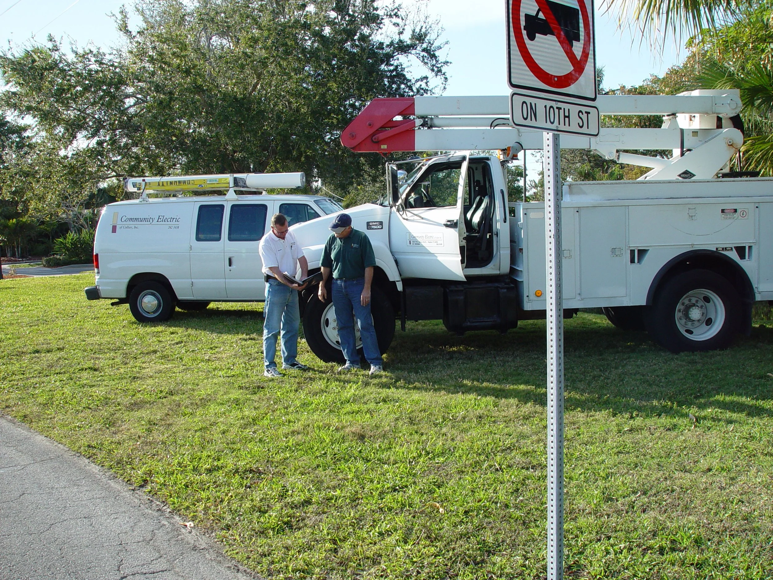 Two workers are standing outside next to a white utility truck, reviewing some documents. There is a street sign indicating no parking on 10th Street, and there are trees and grass in the background.