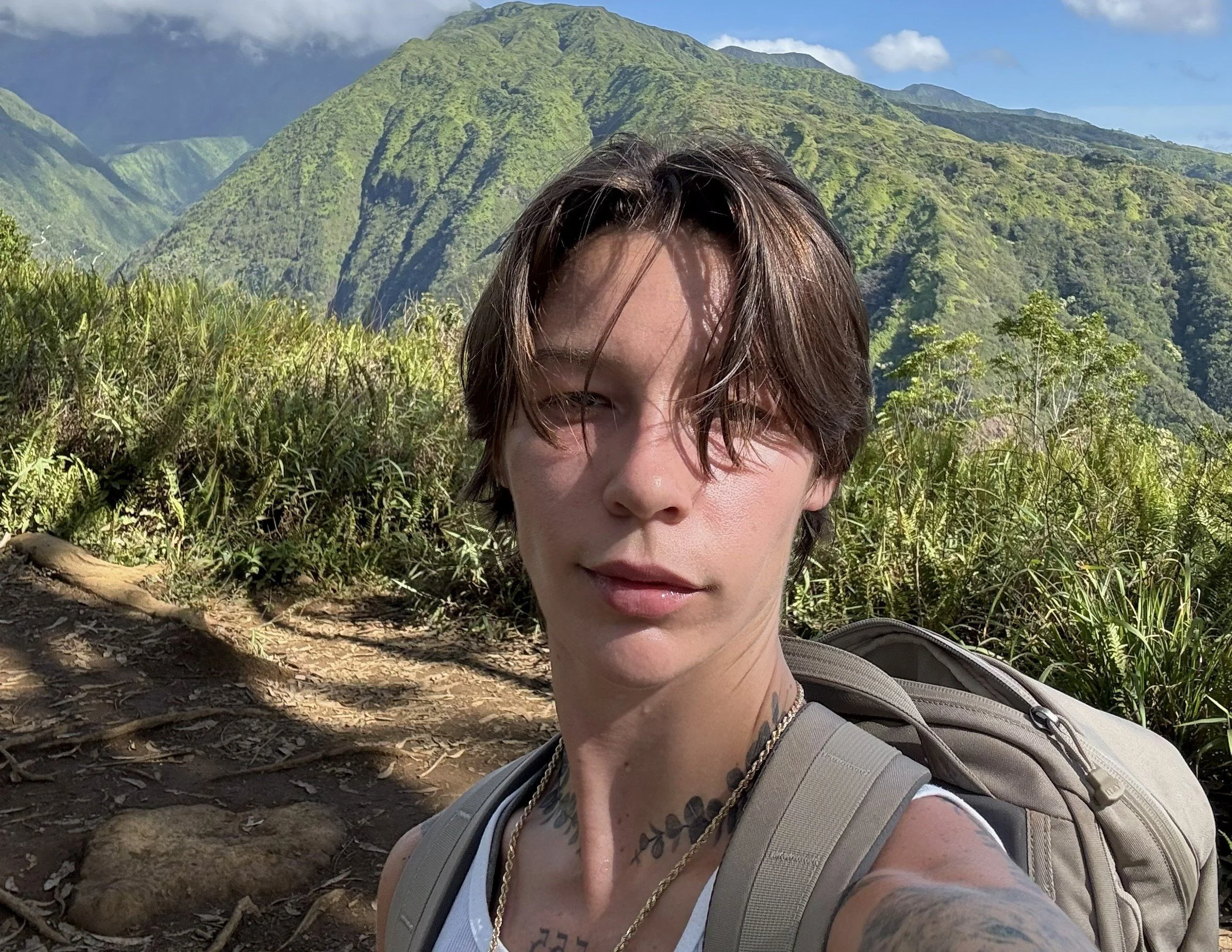 A person with short brown hair and tattoos on their shoulder, wearing a gray backpack and white tank top, taking a selfie on a mountain trail with lush green mountains and a blue sky in the background.