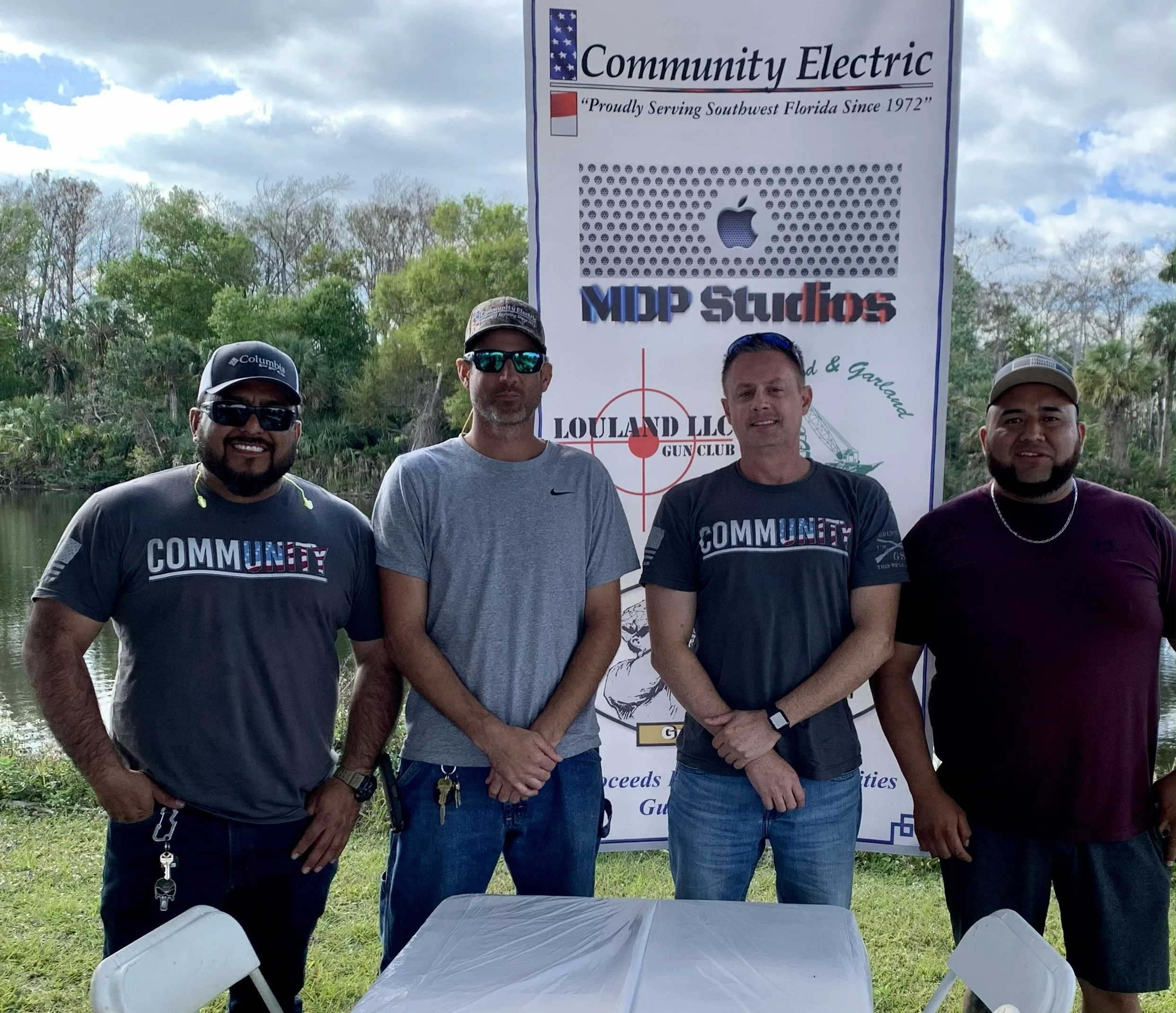 Four men standing outdoors in front of a large banner with logos and text, wearing casual clothing and sunglasses, behind a white table and chairs, with trees and cloudy sky in the background.