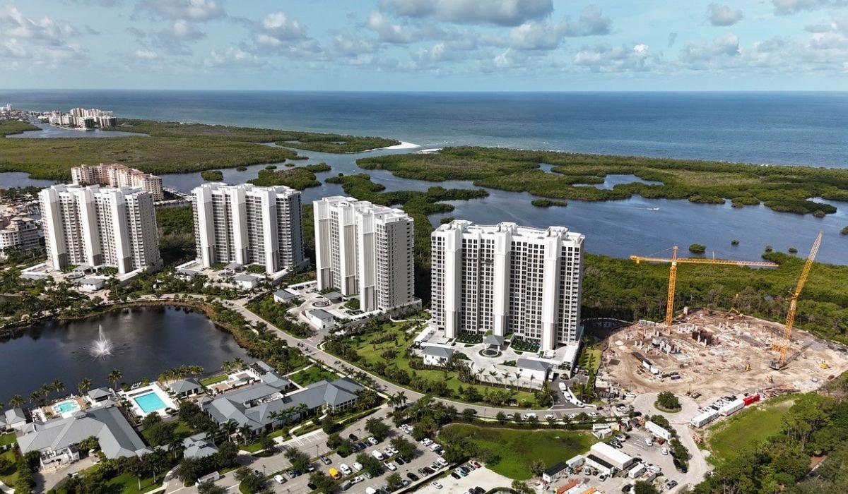 Aerial view of a coastal city with high-rise buildings, water bodies, a construction site with cranes, and the ocean in the background.