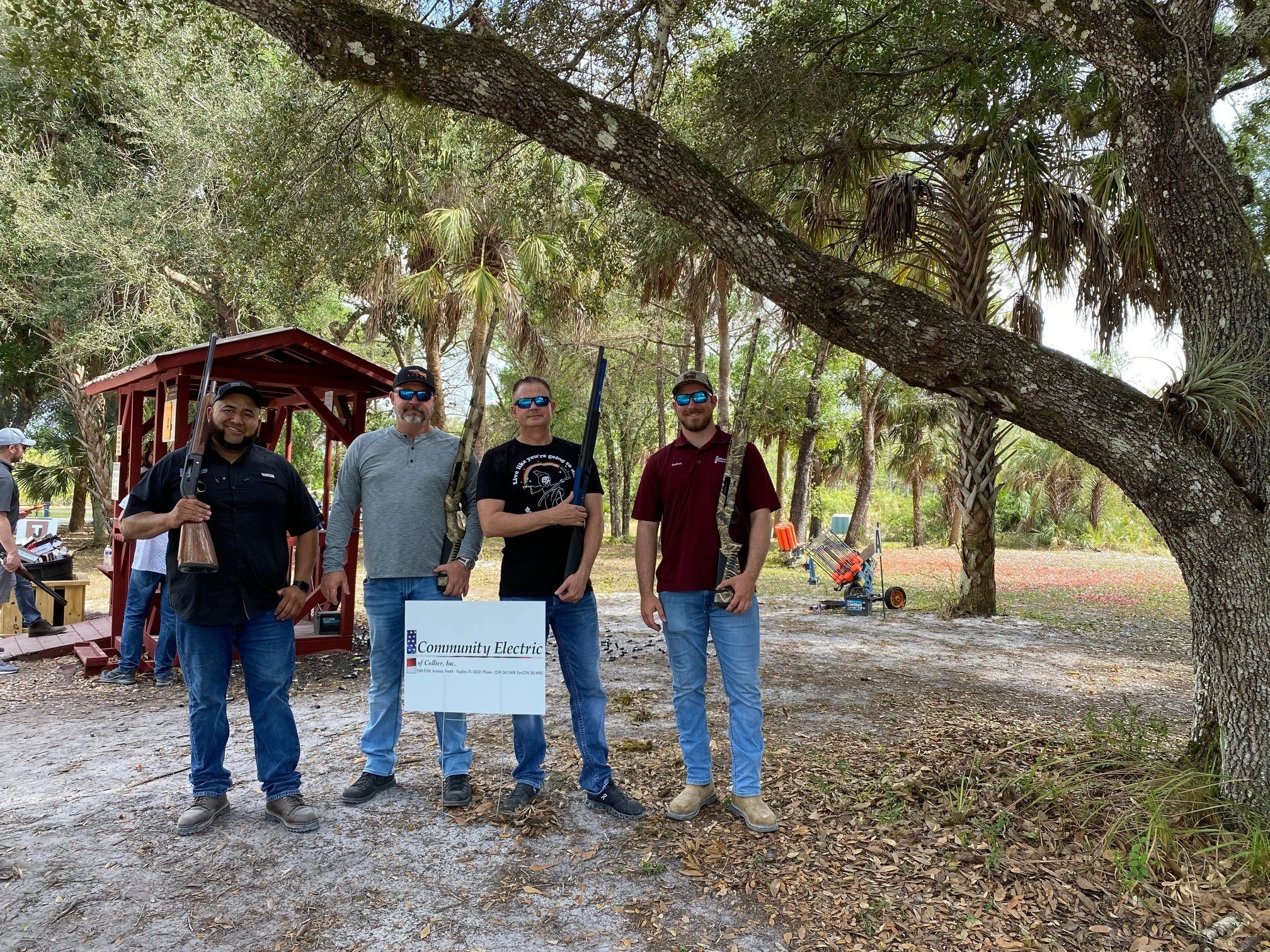 Four men standing outdoors in a wooded area, holding shotguns, in front of a sign that reads "Community Electric." One is wearing sunglasses and a black jacket, the second is wearing sunglasses, a gray shirt, and a gray cap, the third is wearing sung