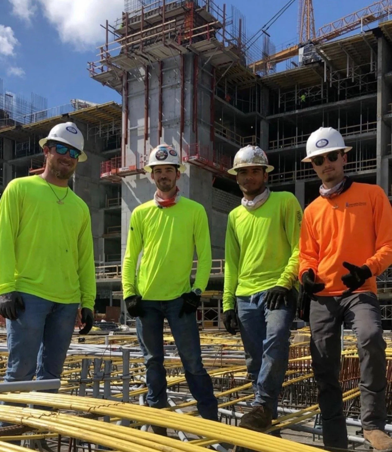 Four construction workers in safety helmets and bright clothing stand in front of Kalea bay in Naples Florida . The workers are smiling at the camera with construction materials and scaffolding behind them.