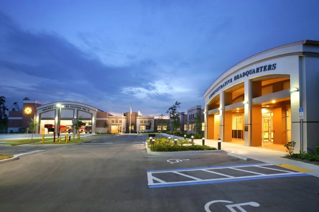 Exterior of a police station or government building at dusk, with parking lot and street lights on.