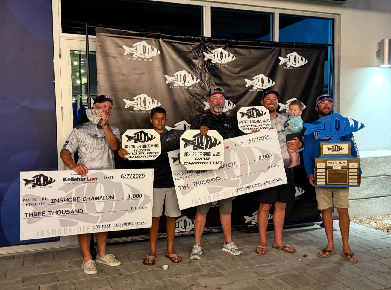 Group of five people standing outdoors at night celebrating a fishing competition victory, holding oversized checks and a trophy shaped like a fish, with a black backdrop featuring a fish logo and the event name 'Inshore Offshore Wars'.