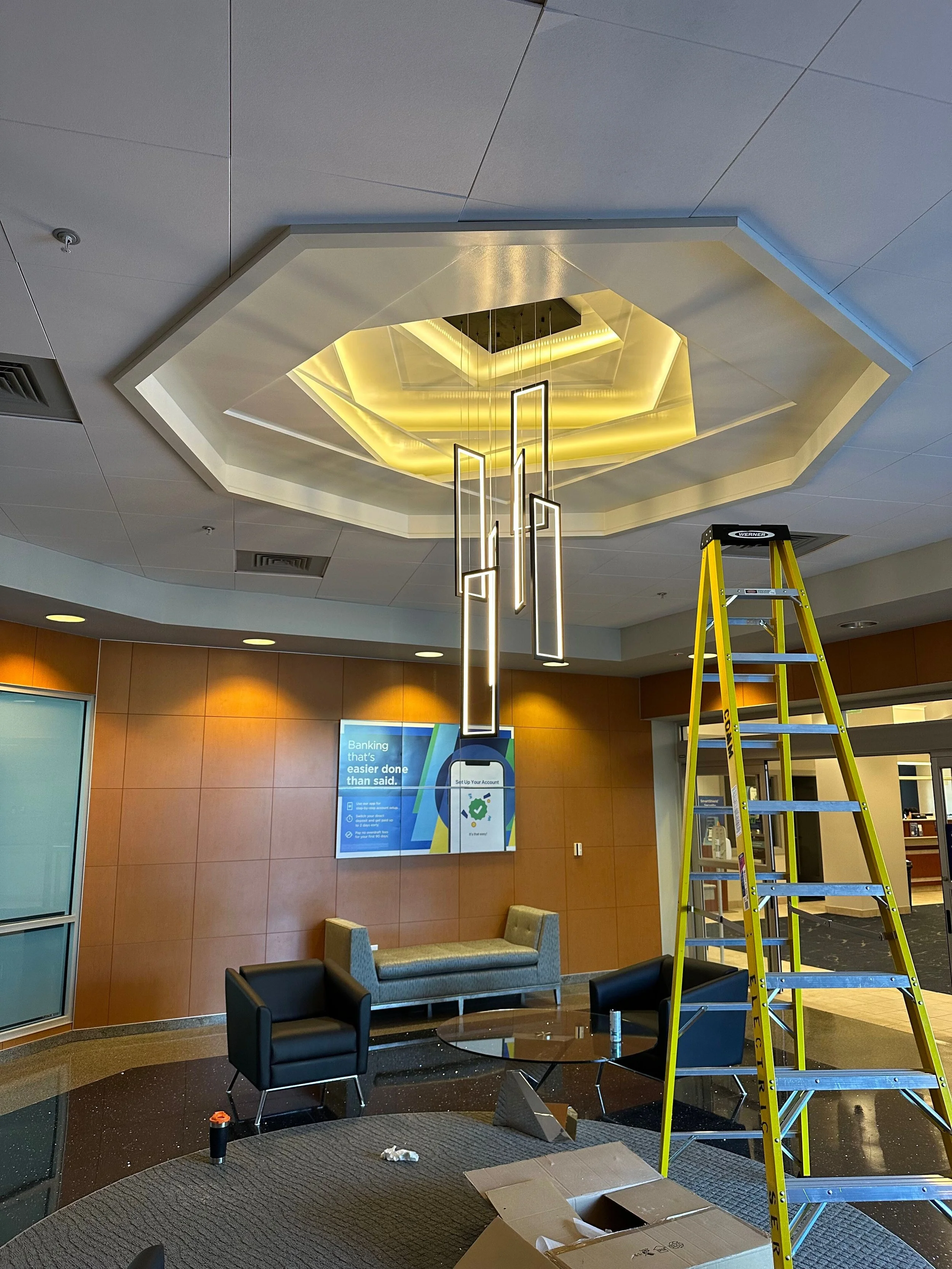 Interior of a modern lobby area with a decorative ceiling, lit hanging light fixtures, a wooden wall with a digital advertising display, and seating areas. A yellow ladder and some boxes are present, indicating ongoing setup or maintenance.