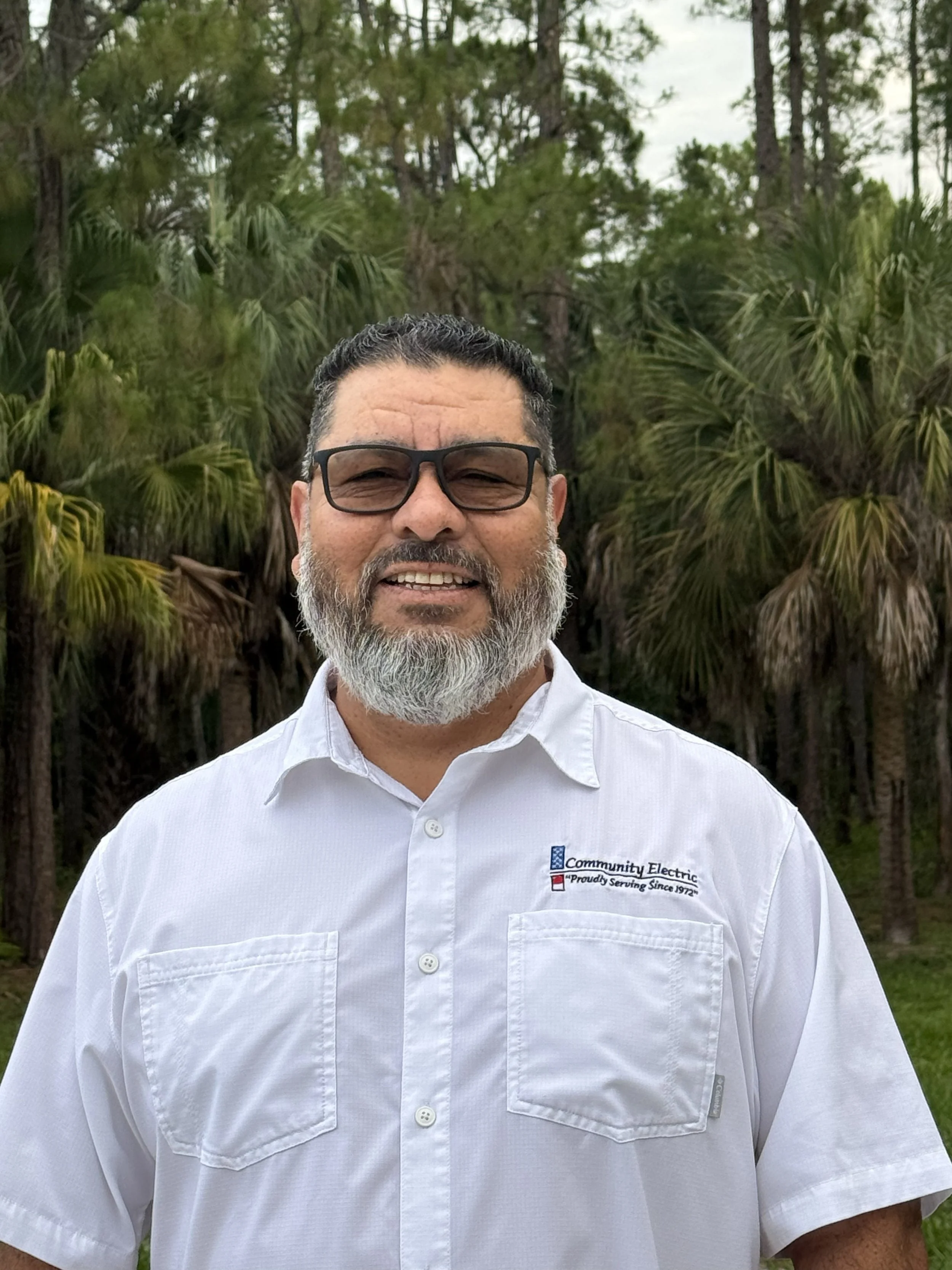 Close-up of a smiling man with a beard and glasses, wearing a white button-up shirt with a logo, standing outdoors with trees in the background.