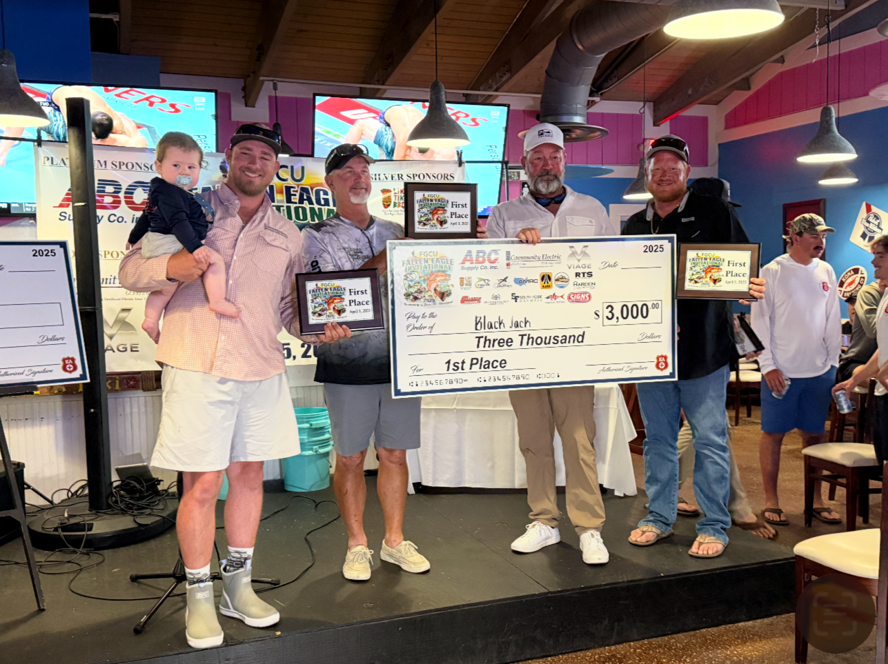 Group of men holding awards and a large check for $3000, celebrating first place at an outdoor event, with a woman holding a young child, at a venue decorated with banners and screens.