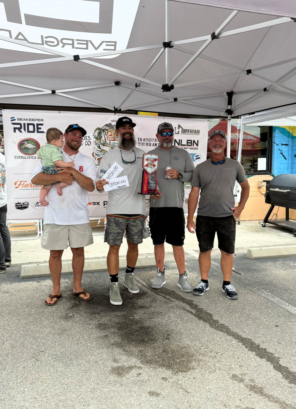 Four men standing under a tent, with one holding a child. One man is holding a trophy and a check, and another has a drink can. All are smiling and posing for a photo at an outdoor event with a banner in the background.