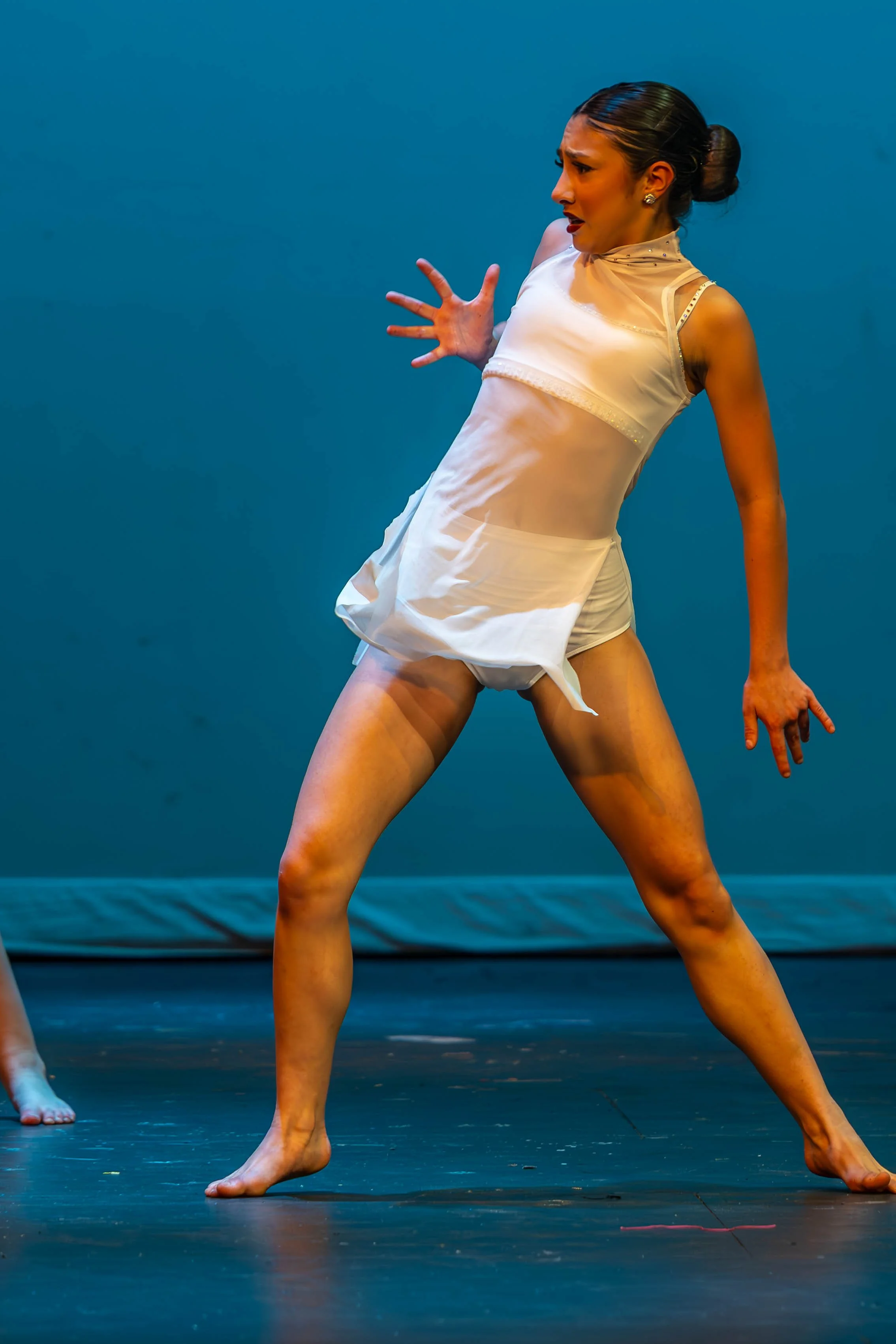 A woman in a white dance costume performing on stage, with her body curved and hands raised in a dance pose, against a plain blue background.