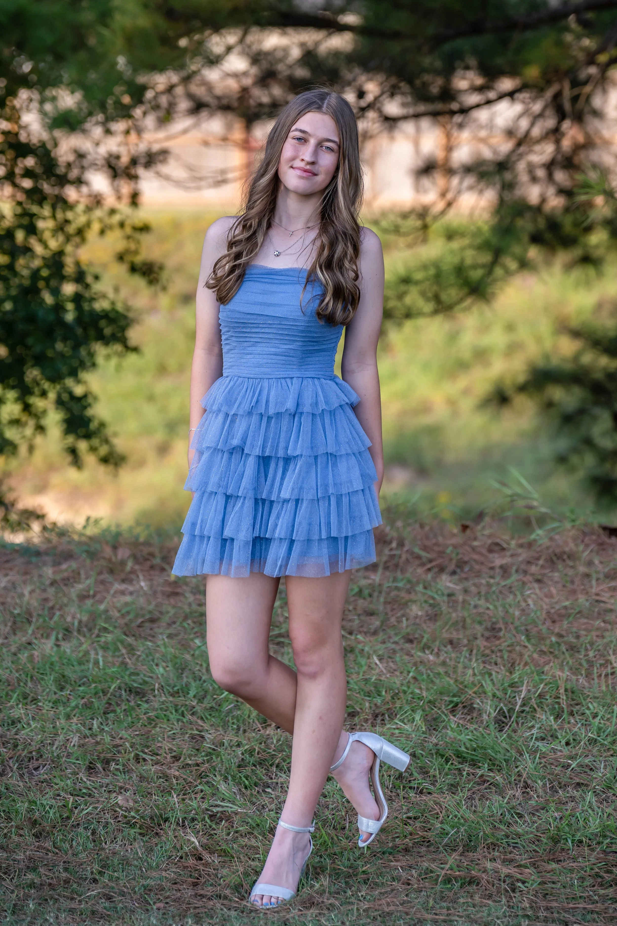 A young woman with long wavy brown hair standing outdoors on grass, wearing a light blue strapless dress with layered ruffles and silver high-heeled sandals, smiling softly at the camera.