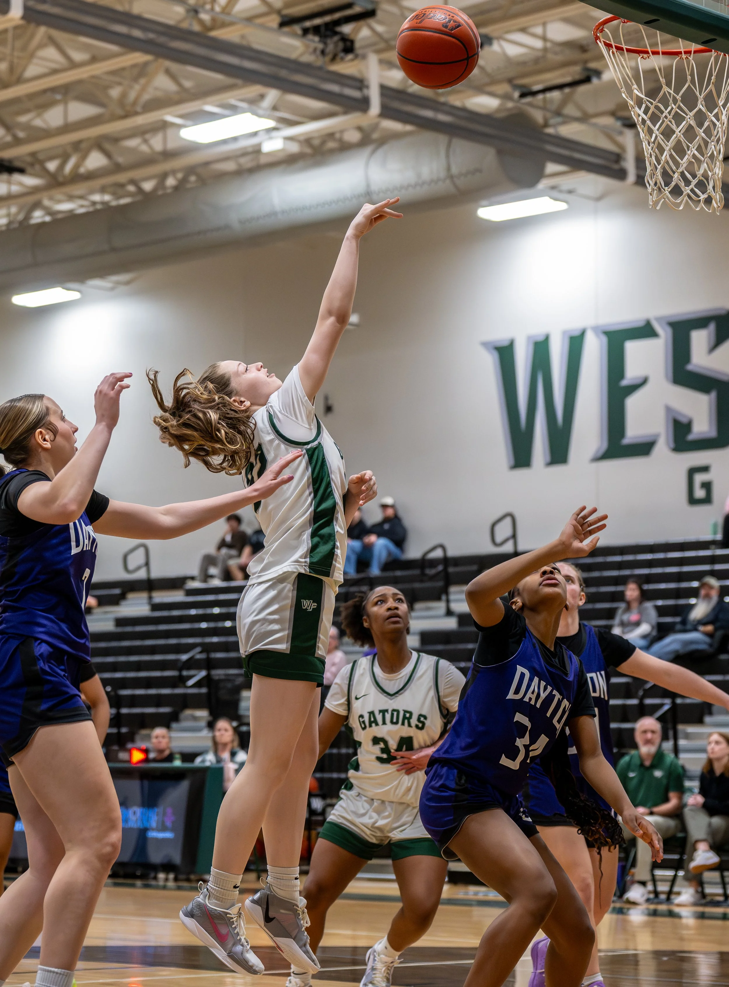 Women playing basketball during a game in an indoor gym, jumping for the ball near the basket.