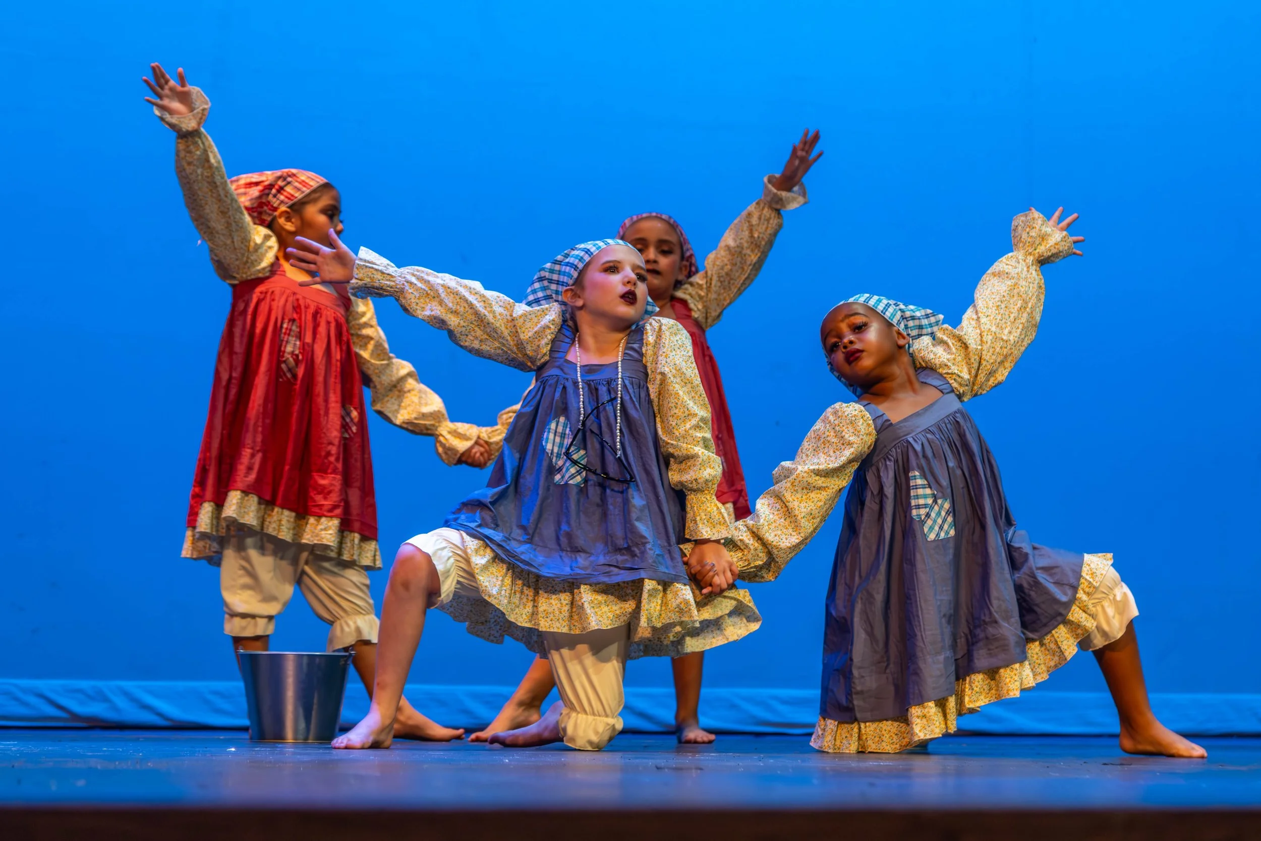 Children dressed in colorful, vintage-style costumes performing a dance on stage under a blue background.