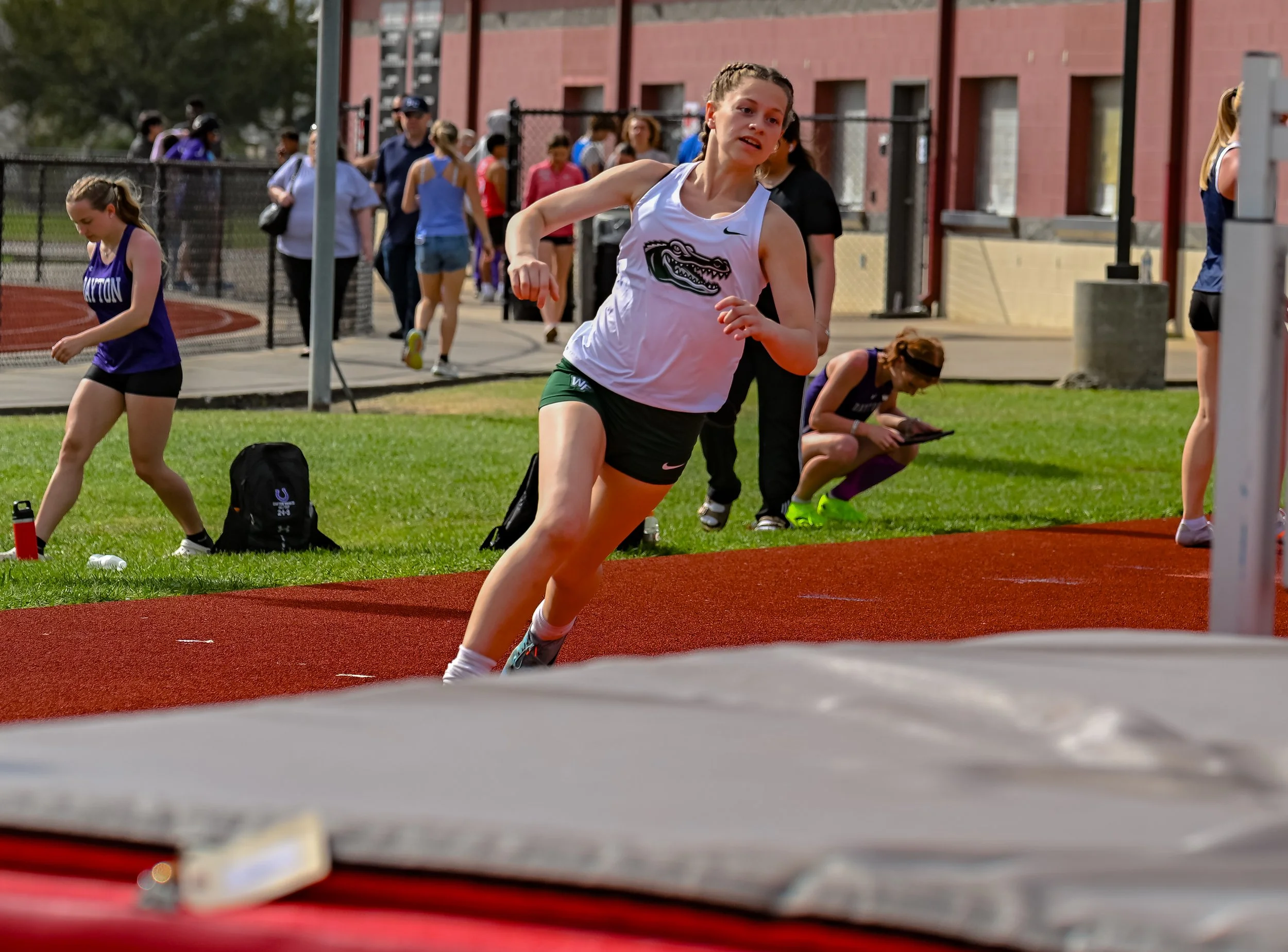 A female athlete in a white and green sports uniform running on a track during a race, with spectators and other runners in the background.