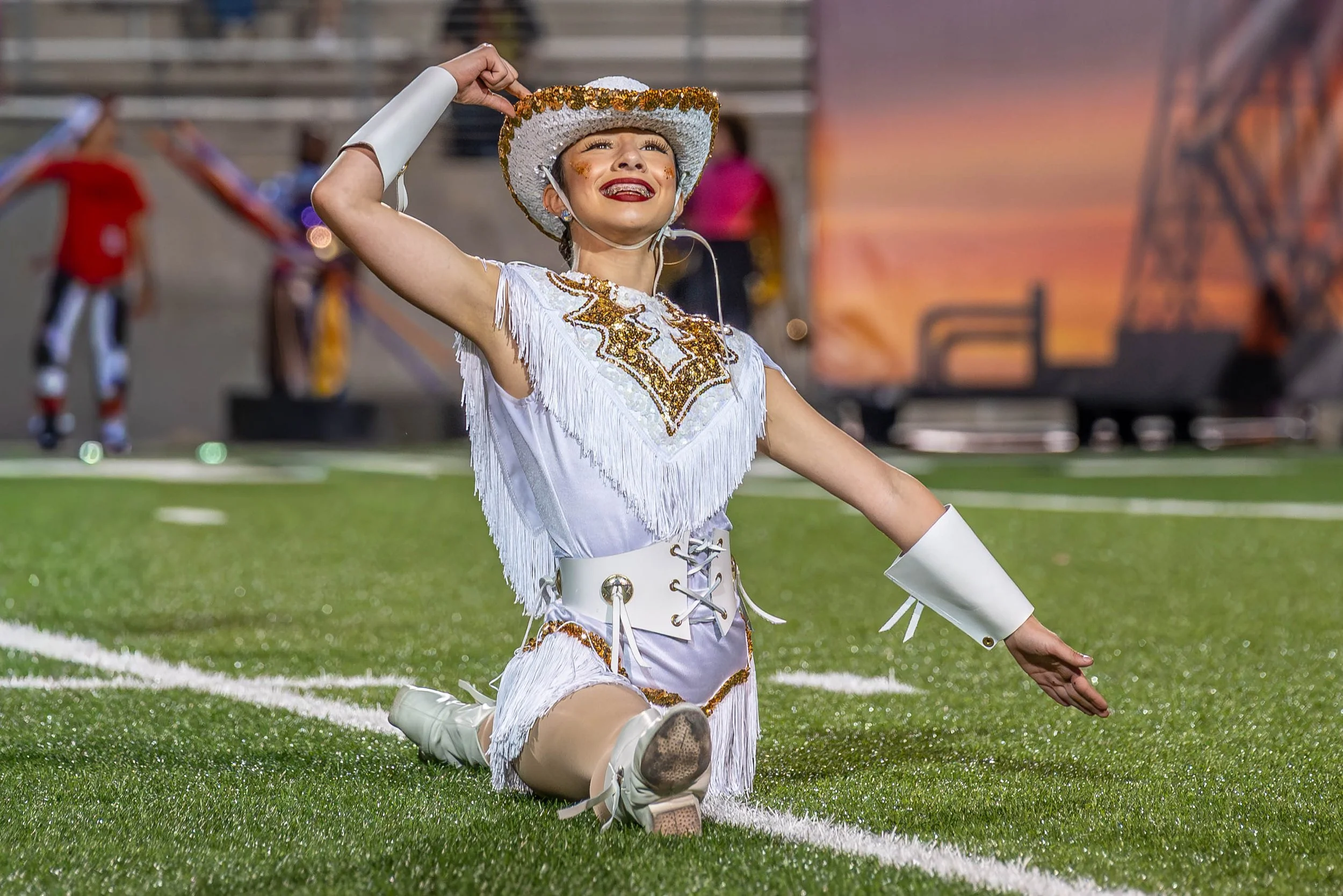 A performer in a colorful costume with gold and white details, kneeling on the field, smiling, with one hand raised to her head and the other extended to the side.