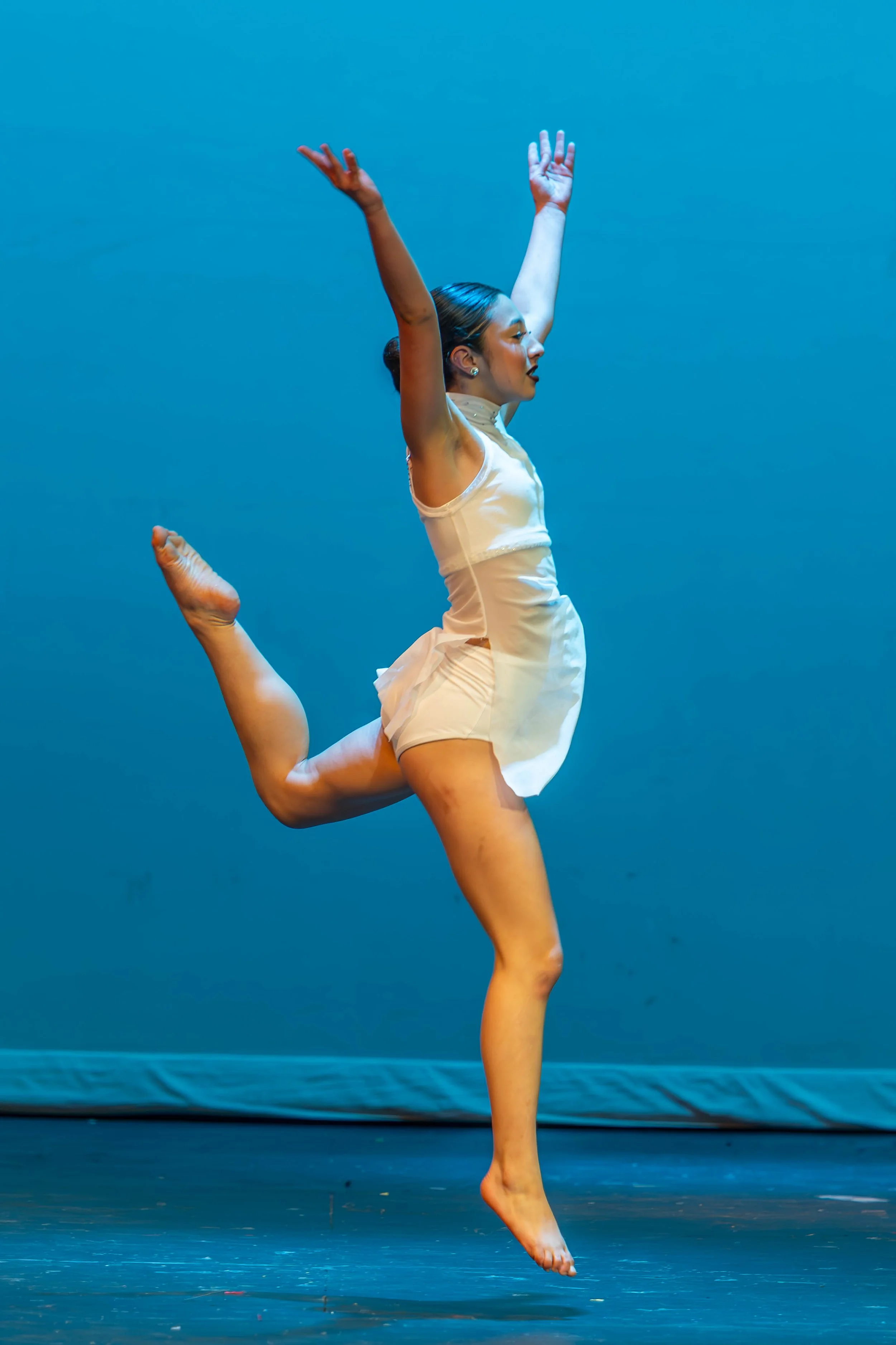 Ballet dancer performing a leap on stage with a blue background, wearing a beige ballet costume and pointe shoes.