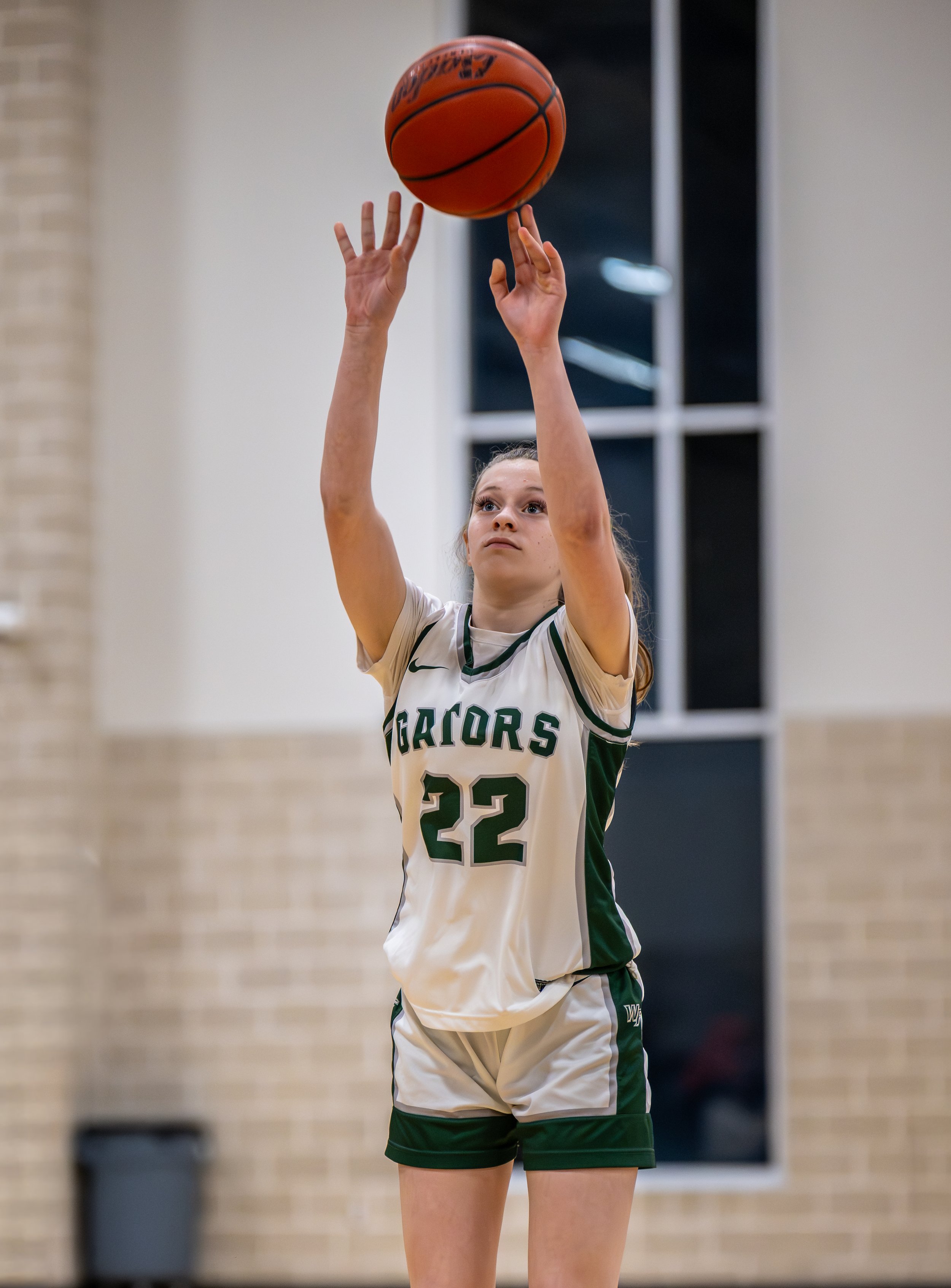 A female volleyball player in a white and green uniform with "GATORS" and the number 22, jumping to hit the ball in an indoor gym.