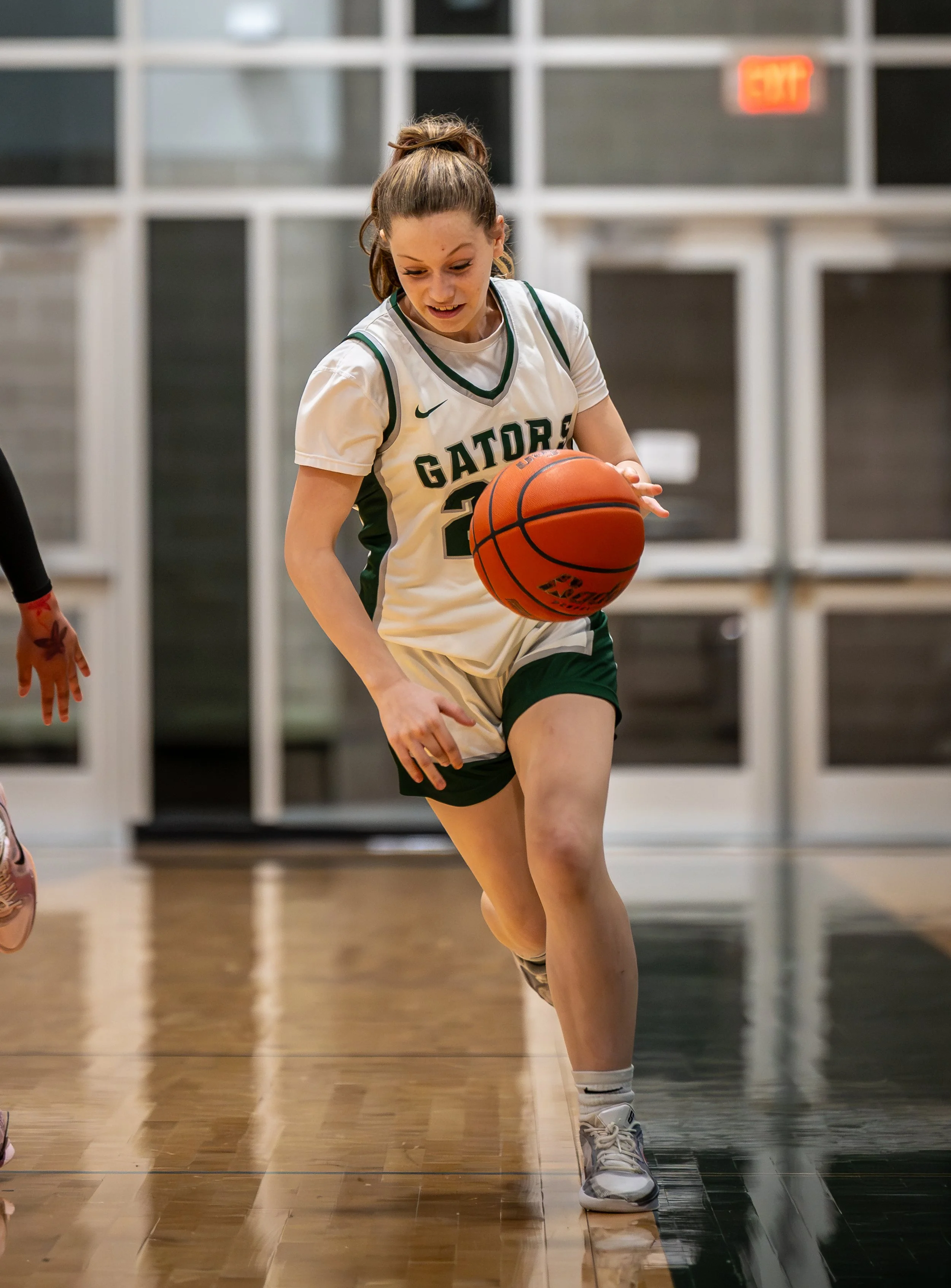 A female basketball player wearing a white uniform with green accents, labeled 'GATORS,' dribbling an orange basketball on an indoor court.