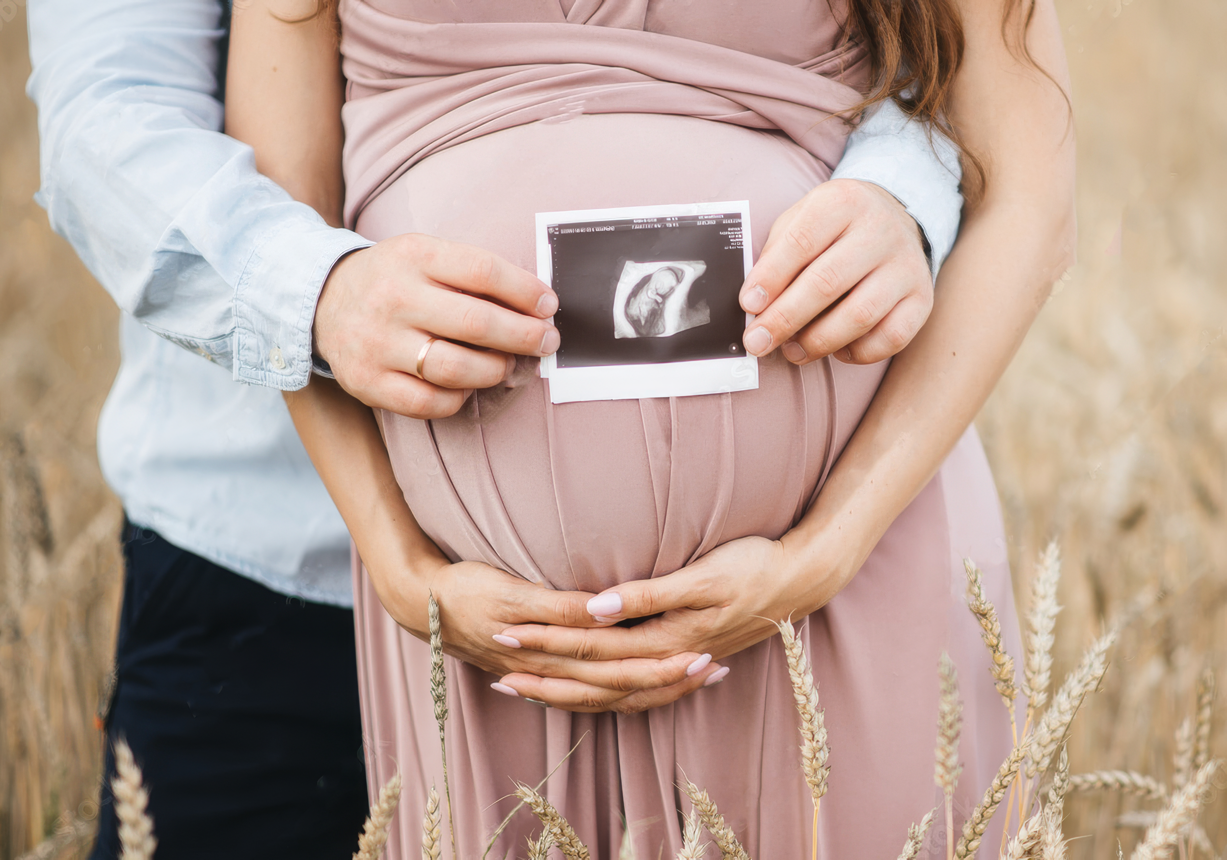 Pregnant woman in pink dress holding ultrasound picture, partner's hands on her stomach, outdoors in grassy area.