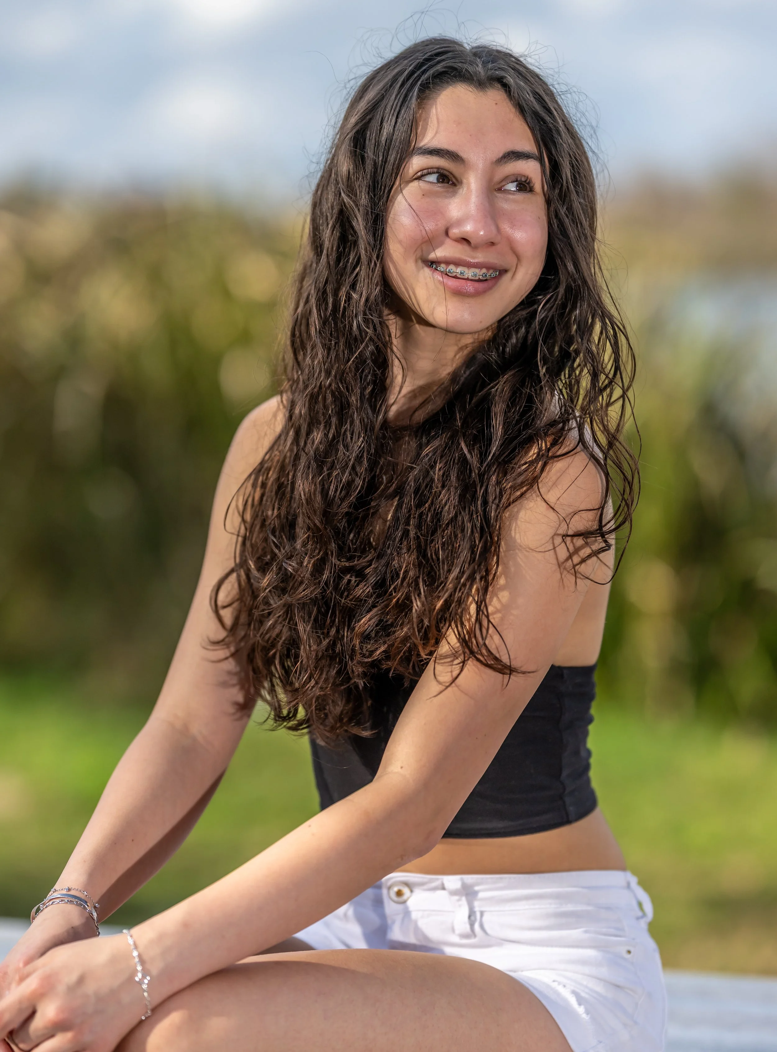 A young woman with long, wavy dark hair smiling and looking to the side, wearing braces, a black top, and white shorts, sitting outdoors with a blurred natural background.