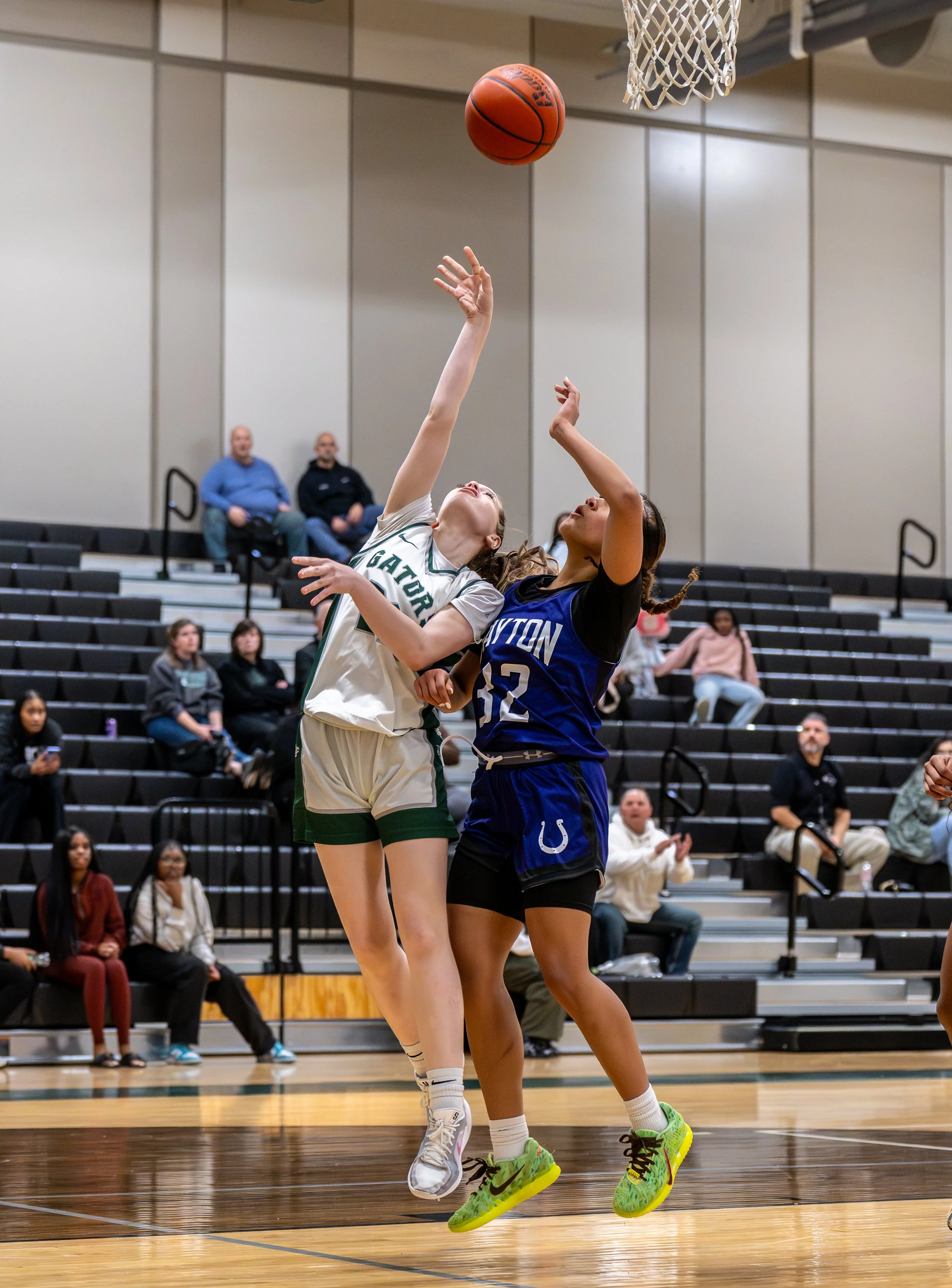 Two female basketball players jump to reach the basketball hoop during a game. One player from the team wearing white and green jerseys and the other from the team wearing blue jerseys. Spectators watch from the bleachers in the background.