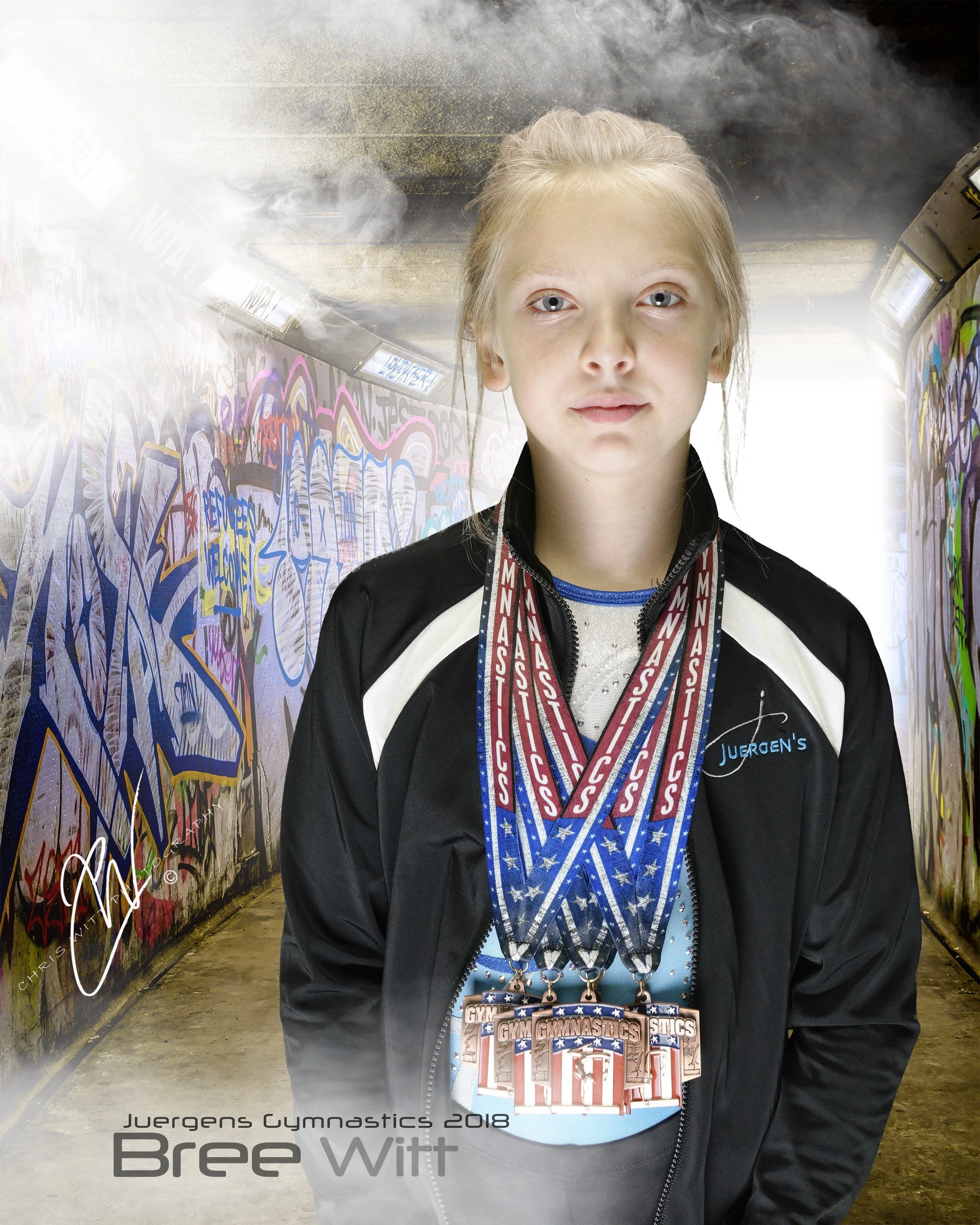 A young gymnast wearing multiple medals around her neck, standing in an indoor setting with graffiti-covered walls and a dark ceiling, holding a large medallion, with the text 'Juergens Gymnastics 2018 Bree Witt' visible.