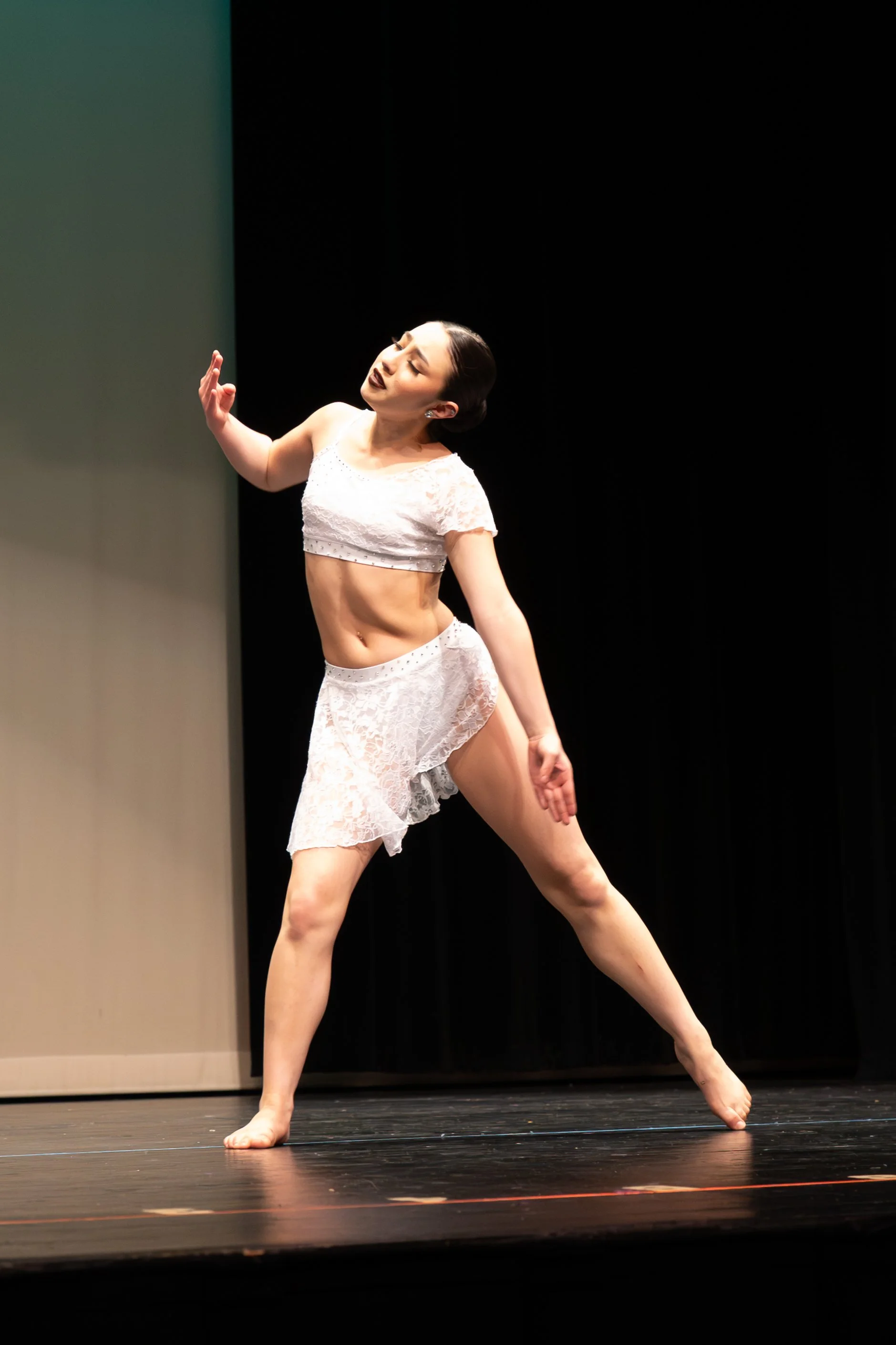 A female dancer in a white lace costume performing on stage, balancing on one foot with her other leg extended.