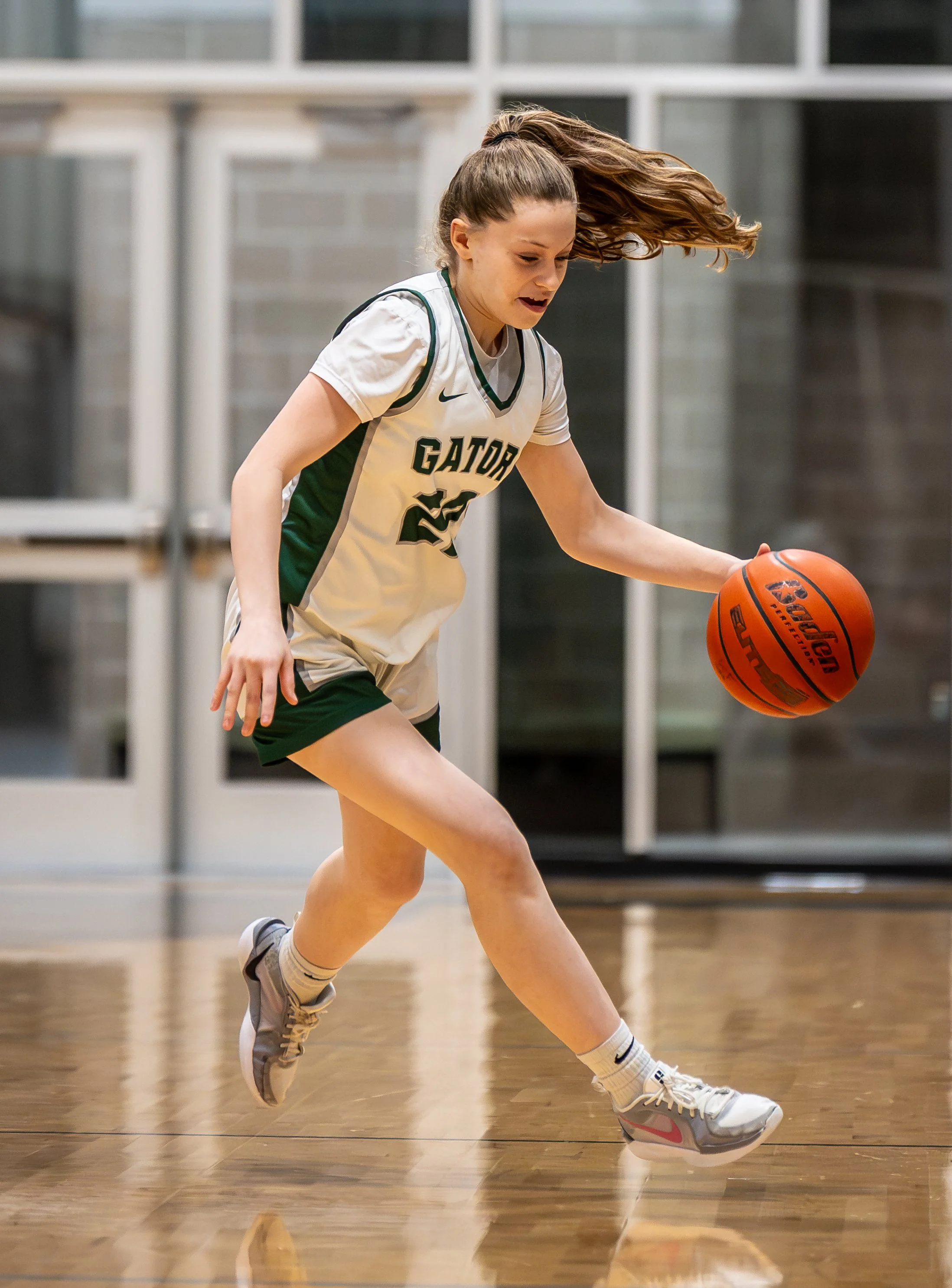 A young female basketball player in a white and green Gator uniform dribbling a basketball on an indoor court.