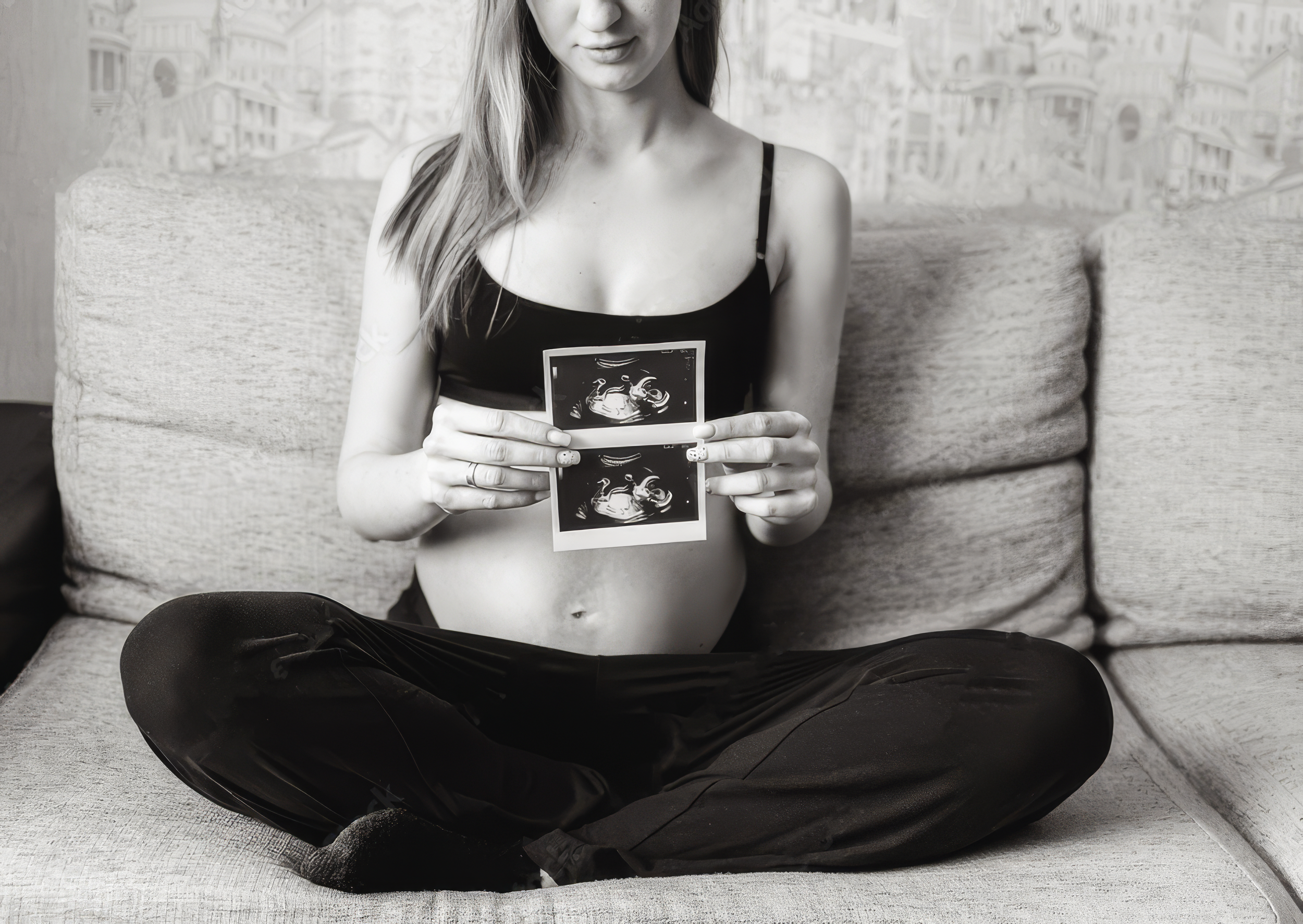 A woman sitting cross-legged on a couch, holding ultrasound images in front of her chest, with a neutral expression, and dressed in a camisole and dark pants. The background has a neutral-colored cushion and a textured wall.
