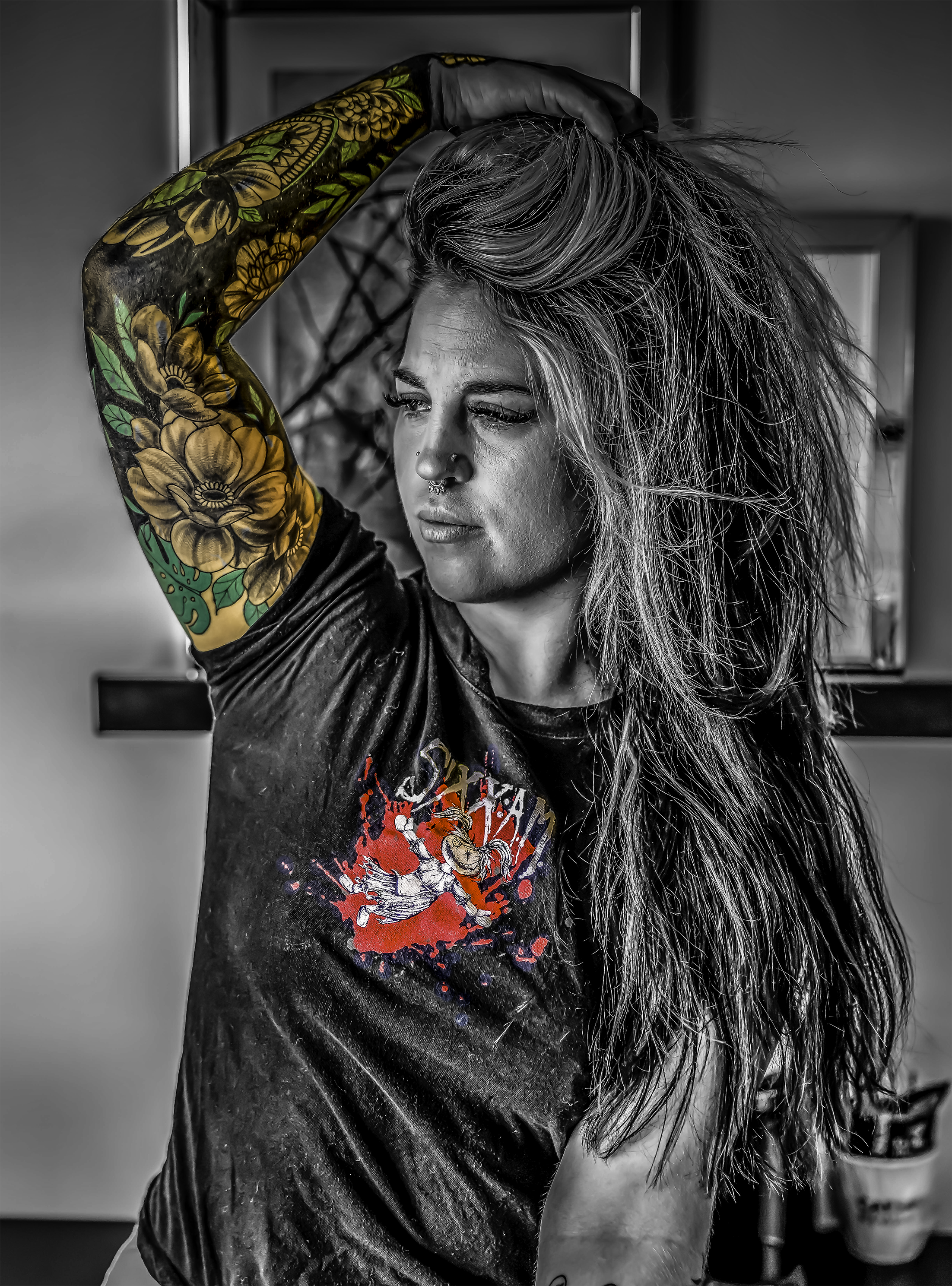 A woman with long, wavy hair in black and white, wearing a graphic T-shirt, is posing with her left arm raised, holding her hair back, revealing a colorful tattoo sleeve on her left arm.