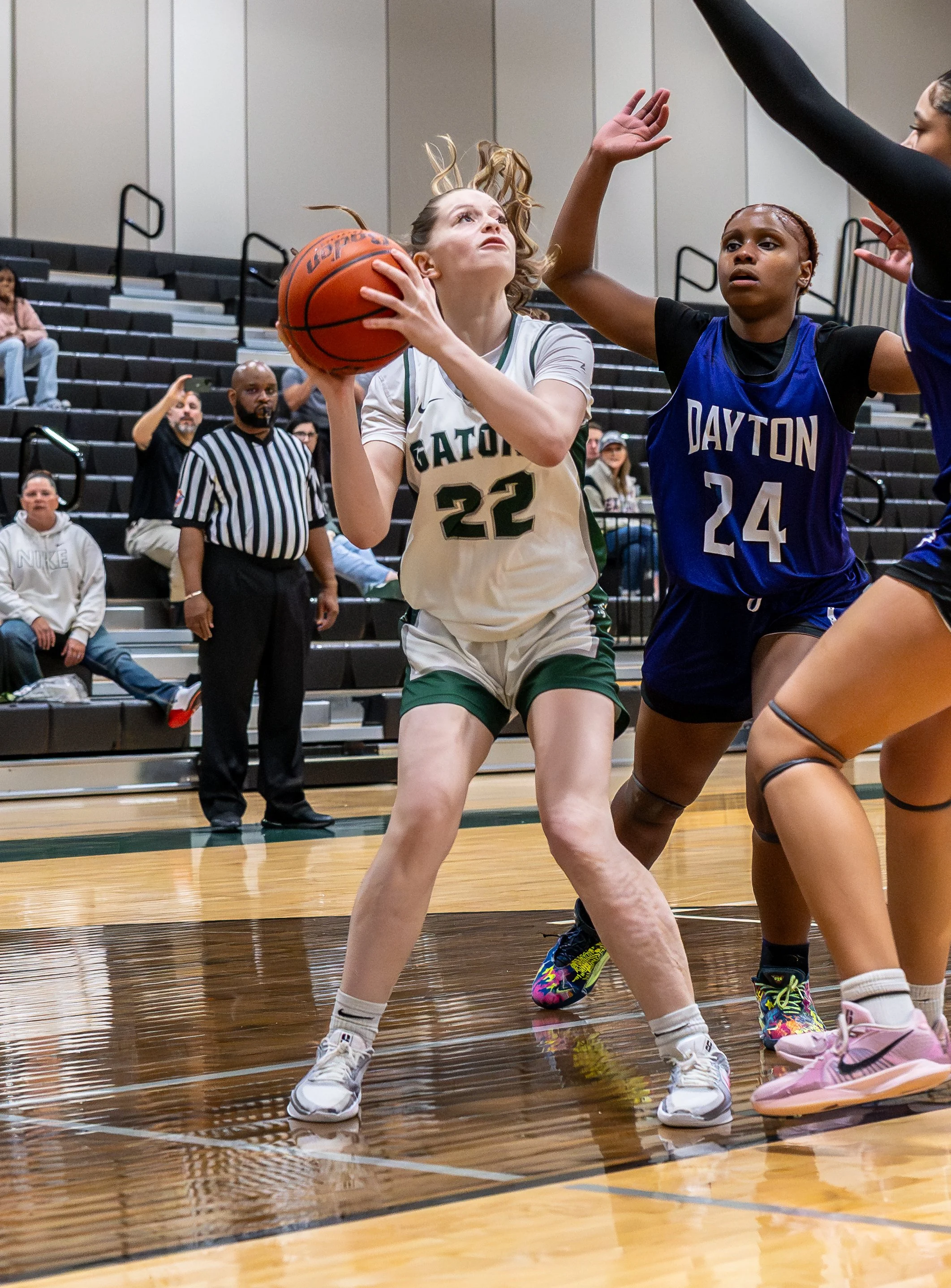 A female basketball player in a white and green uniform holding a basketball during a game against a female player in a blue uniform. The scene takes place on a basketball court with spectators in the background.