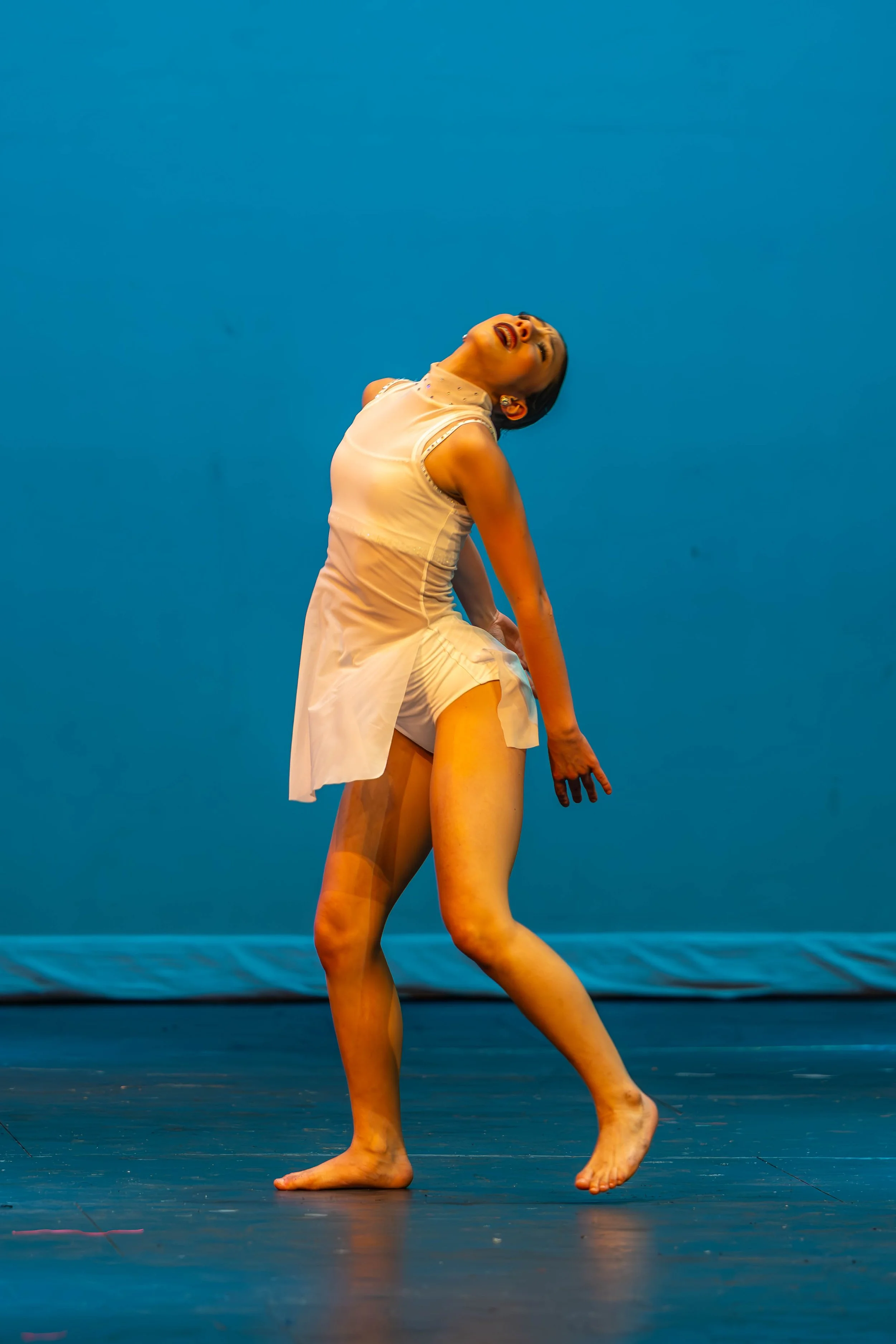 A dancer performing on stage, wearing a sleeveless white dress, appears to be in an expressive moment with her head tilted back and eyes closed, set against a solid blue background.