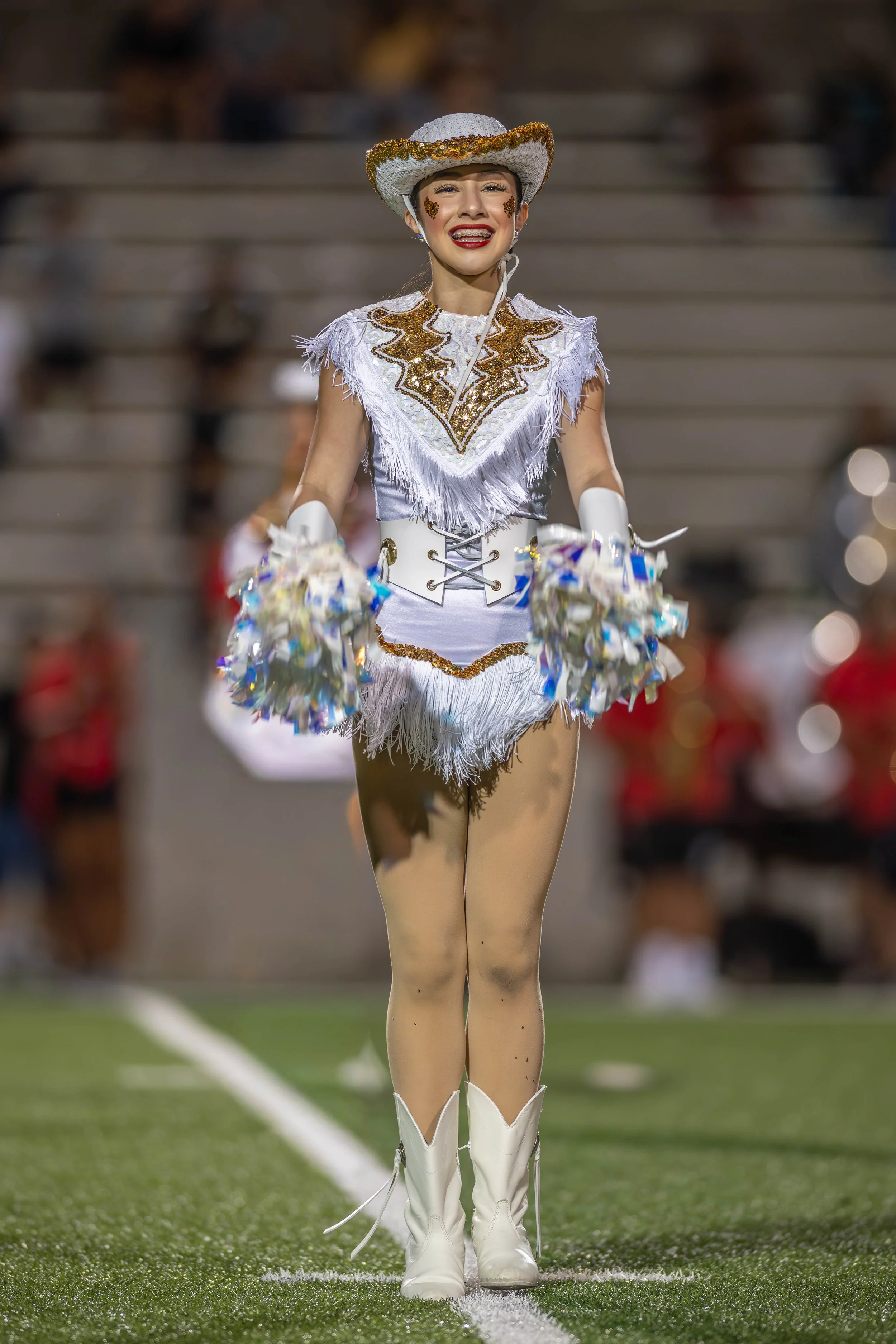 A cheerleader on a football field during a game, wearing a white and gold costume with fringes, cowboy hat, and holding pom-poms.