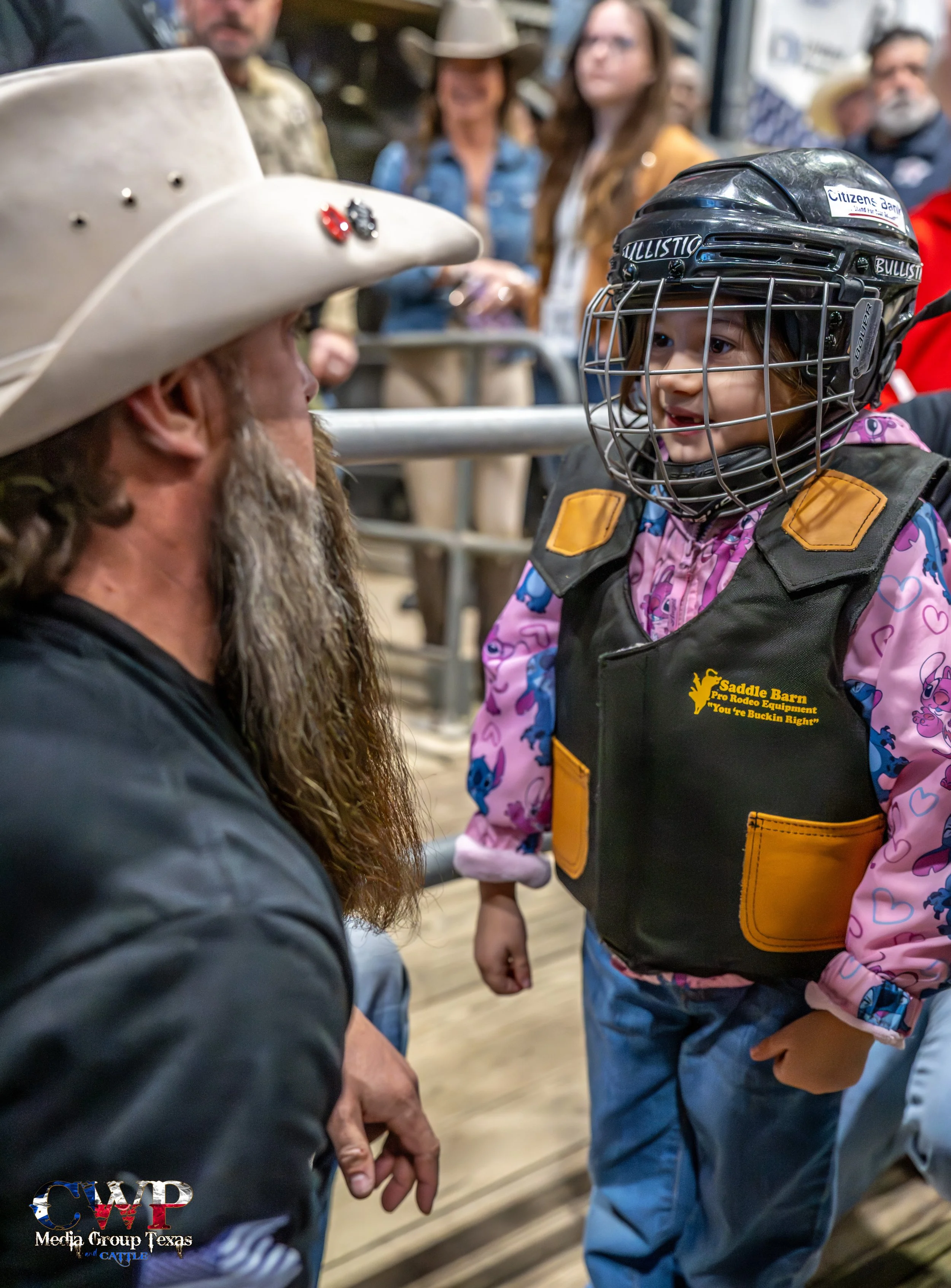 A young girl wearing a hockey helmet with a wire cage, a vest that says 'Saddle Barn Pro Rodeo Equipment' with a slogan underneath, and a jacket with a colorful pattern, is smiling and looking at an older man with a beard and long hair wearing a cowboy hat and black clothing. They are in a rodeo or similar arena with a crowd of people in the background.
