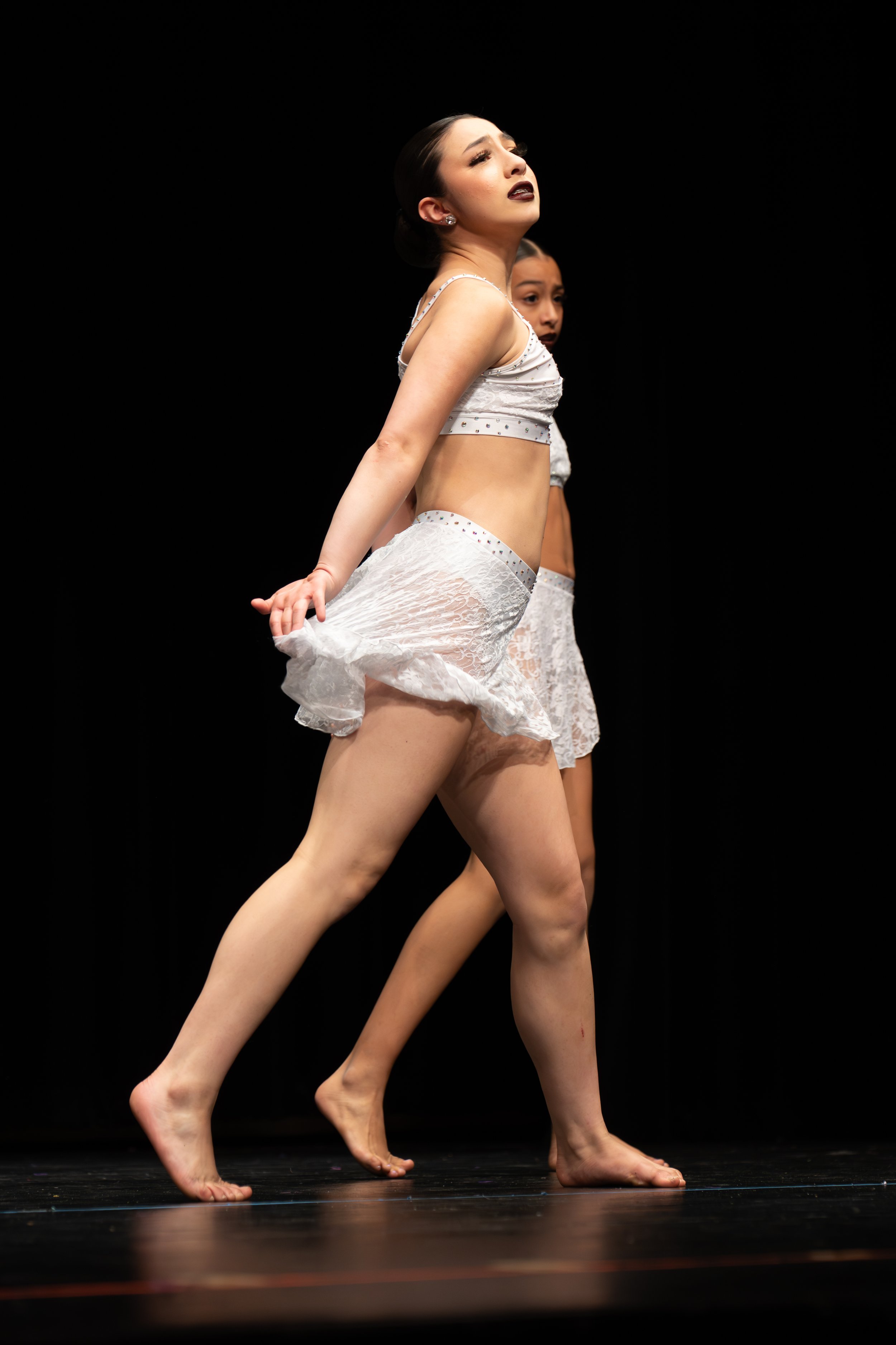 Two female dancers are performing on stage with a black background, dressed in white costumes with lace and rhinestones, captured mid-movement with one leg forward and hands lifting the skirt.