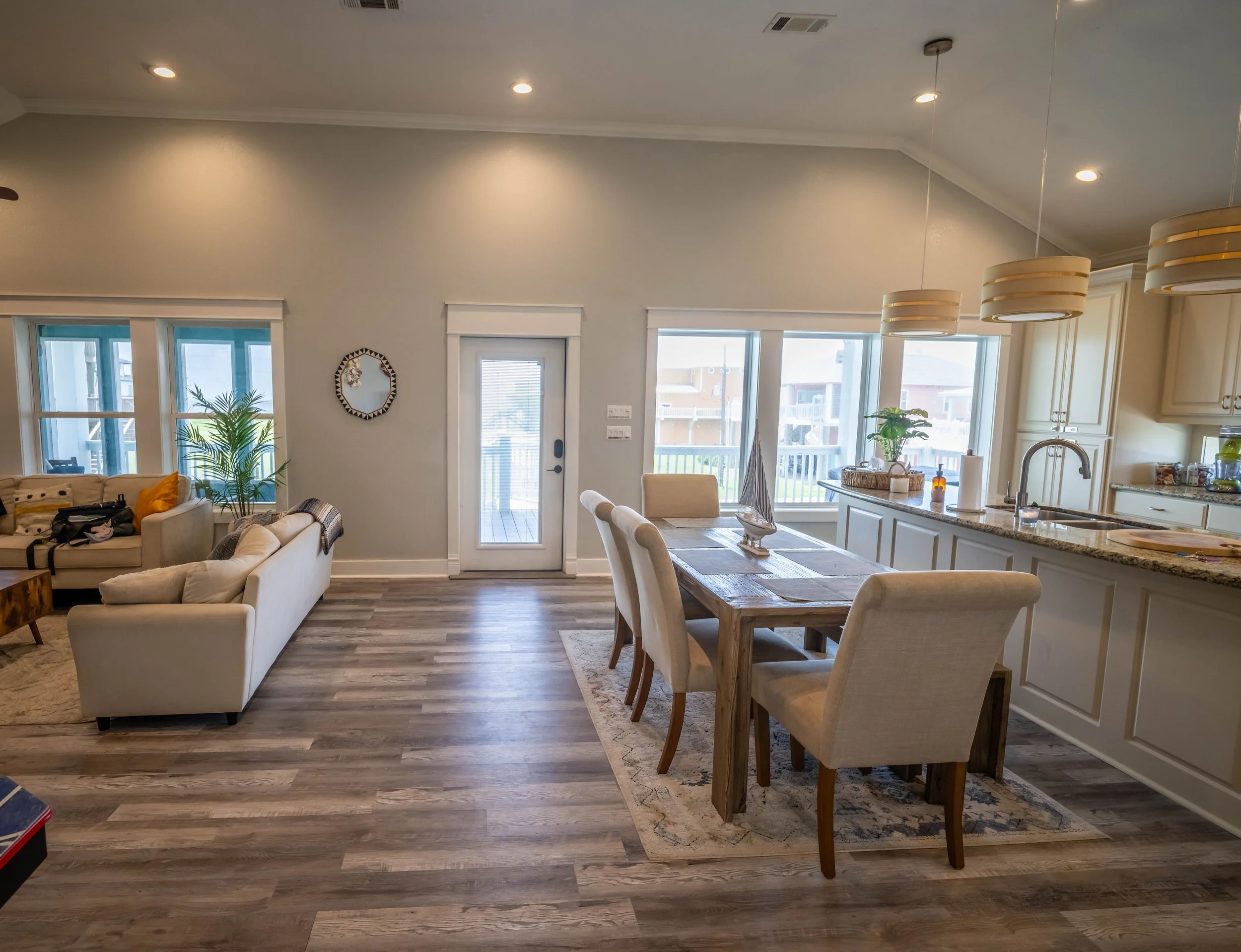 Interior view of a modern, open-plan living and dining area with large windows, a kitchen counter, and seating. Light-colored walls and wood flooring are visible, with a plant near the windows and hung pendant lights.
