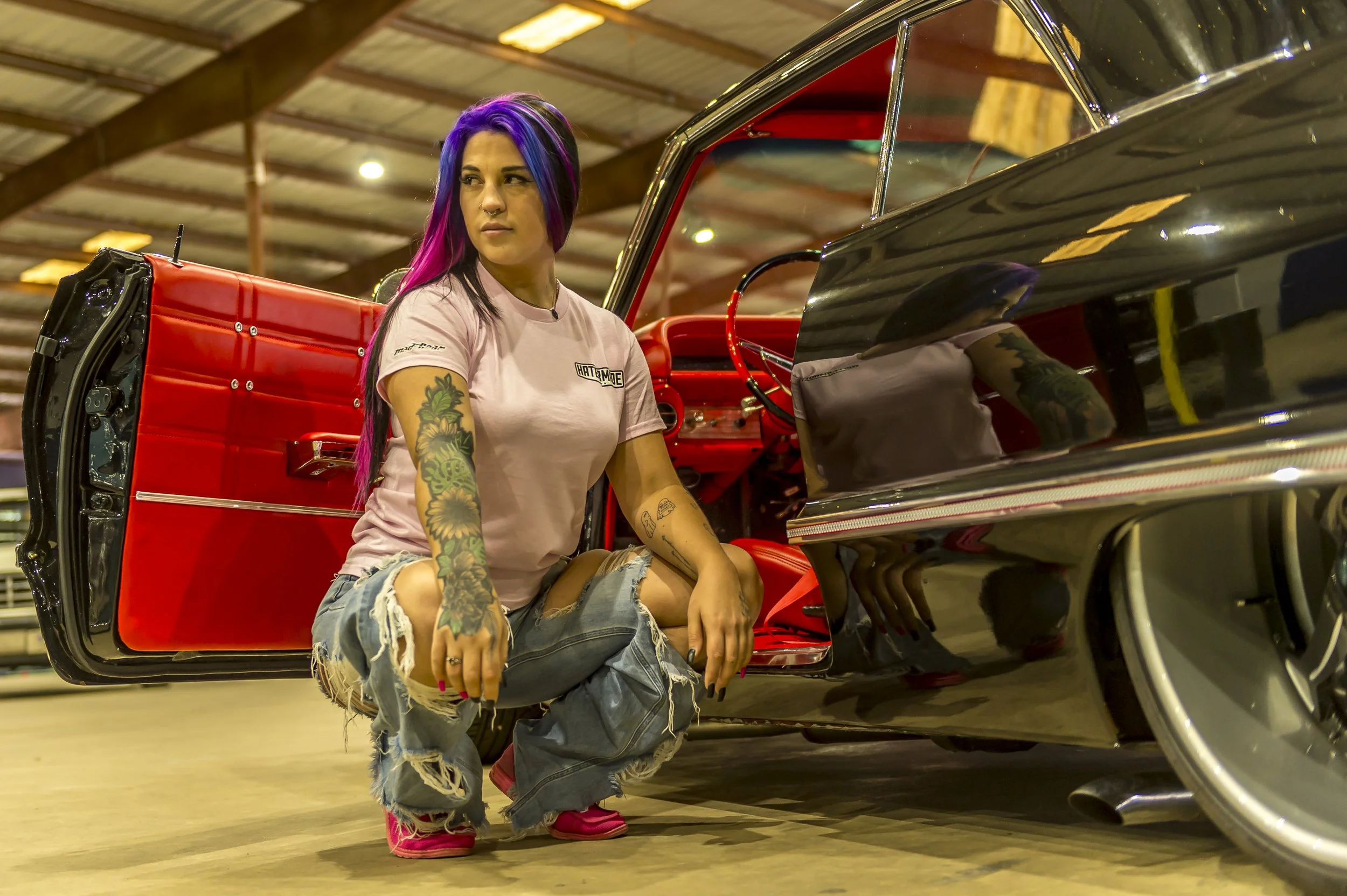 A woman with purple and pink hair sitting beside a vintage black and red car inside a warehouse.