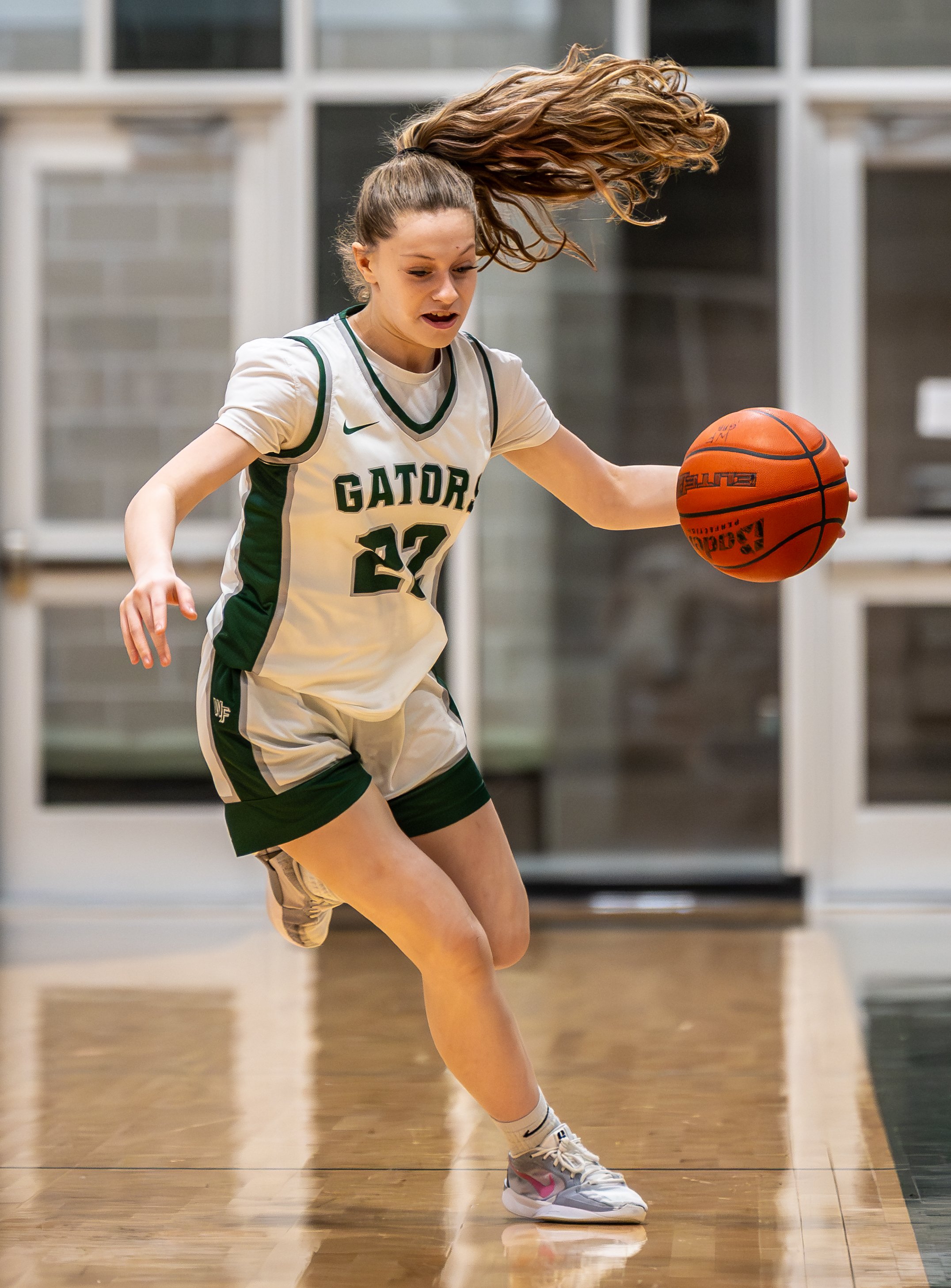 A young female basketball player in a white and green uniform dribbling the basketball on a court, with her hair flying in the air.