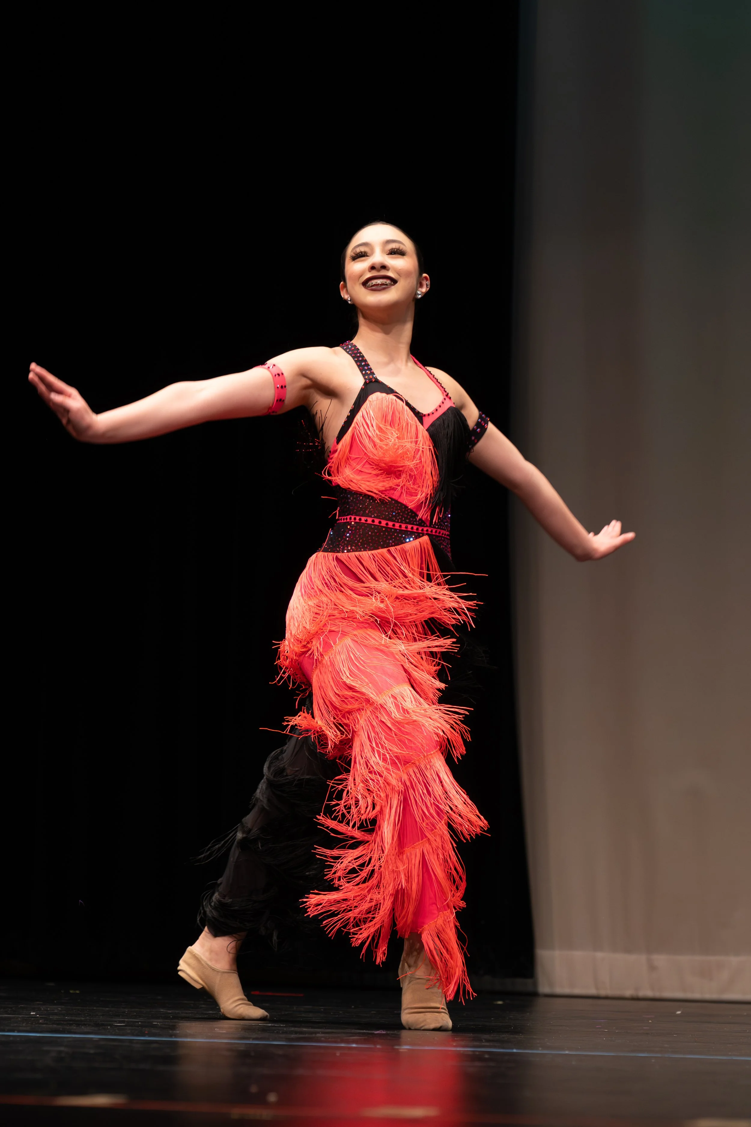A woman performing a dance on stage wearing a vibrant orange dress with fringe details, black accents, and beige dance shoes, against a dark background.