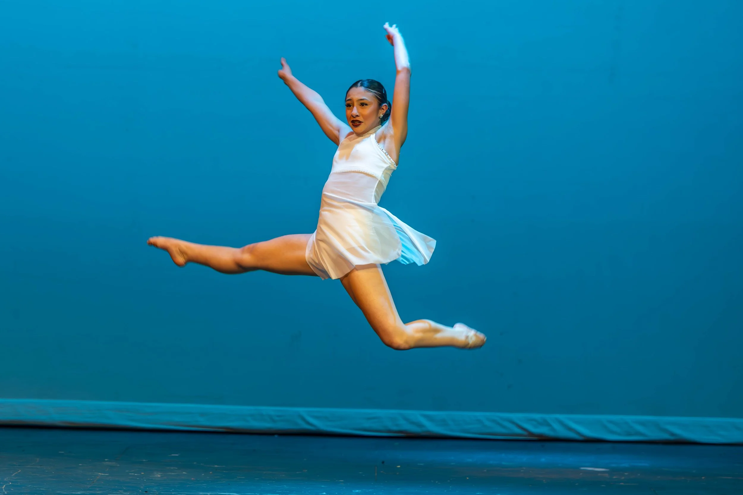 A female dancer in a white dress performing an elegant leap on stage with a plain blue backdrop.