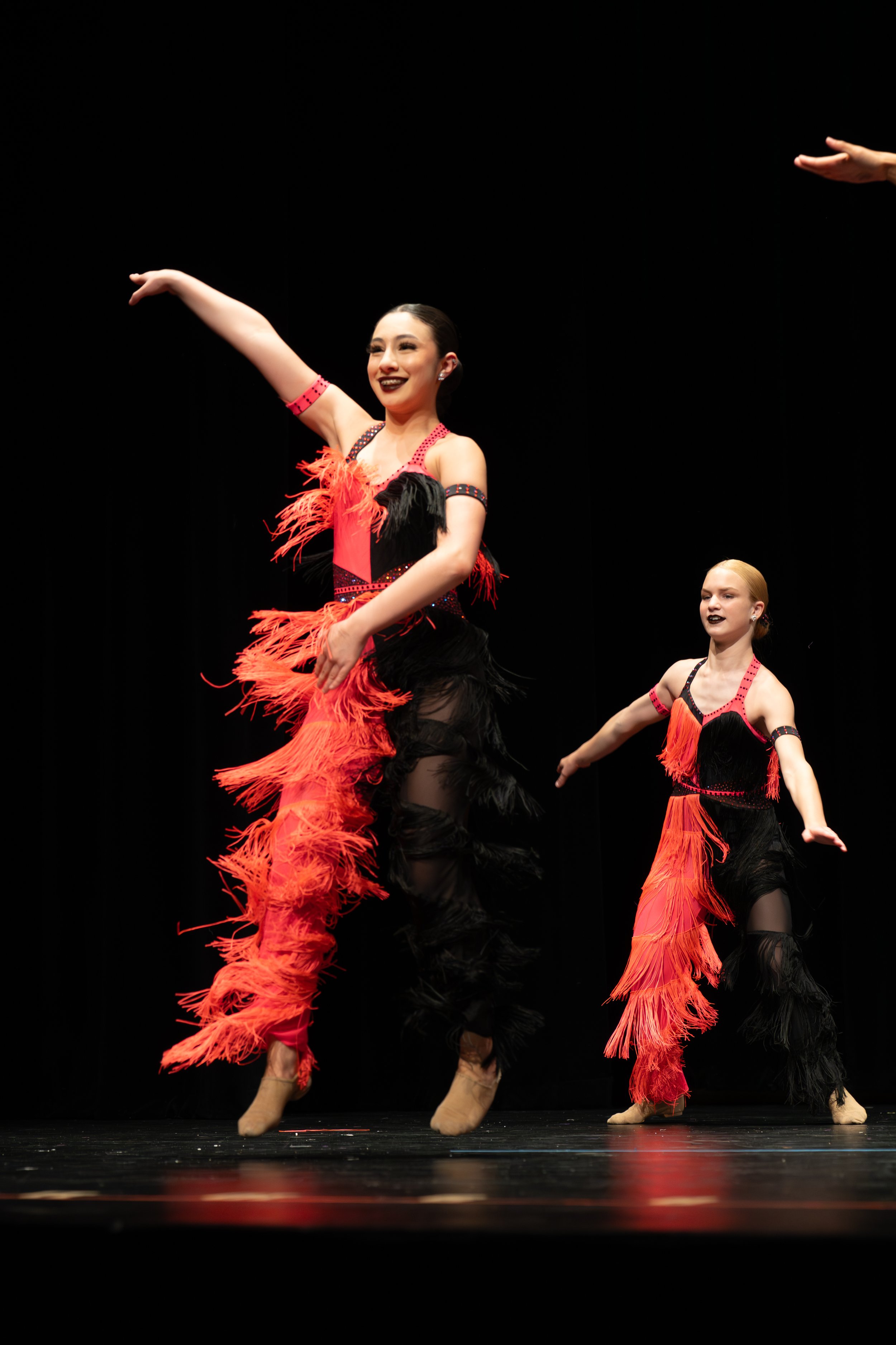Two female dancers perform on stage in colorful costumes with feathered skirts, one in a black and red outfit and the other in a matching design, against a black background.
