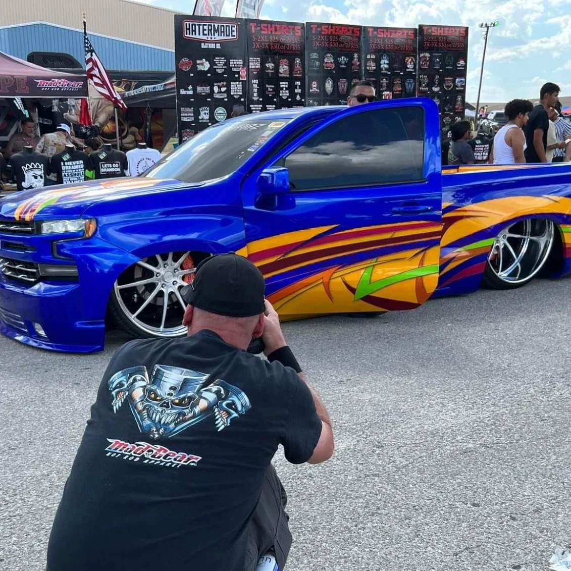 A lowrider truck with vibrant blue paint and colorful flame graphics, parked at a car show. A man in a black cap and graphic t-shirt is taking a photo of the truck, with a crowd and vendor booths in the background.