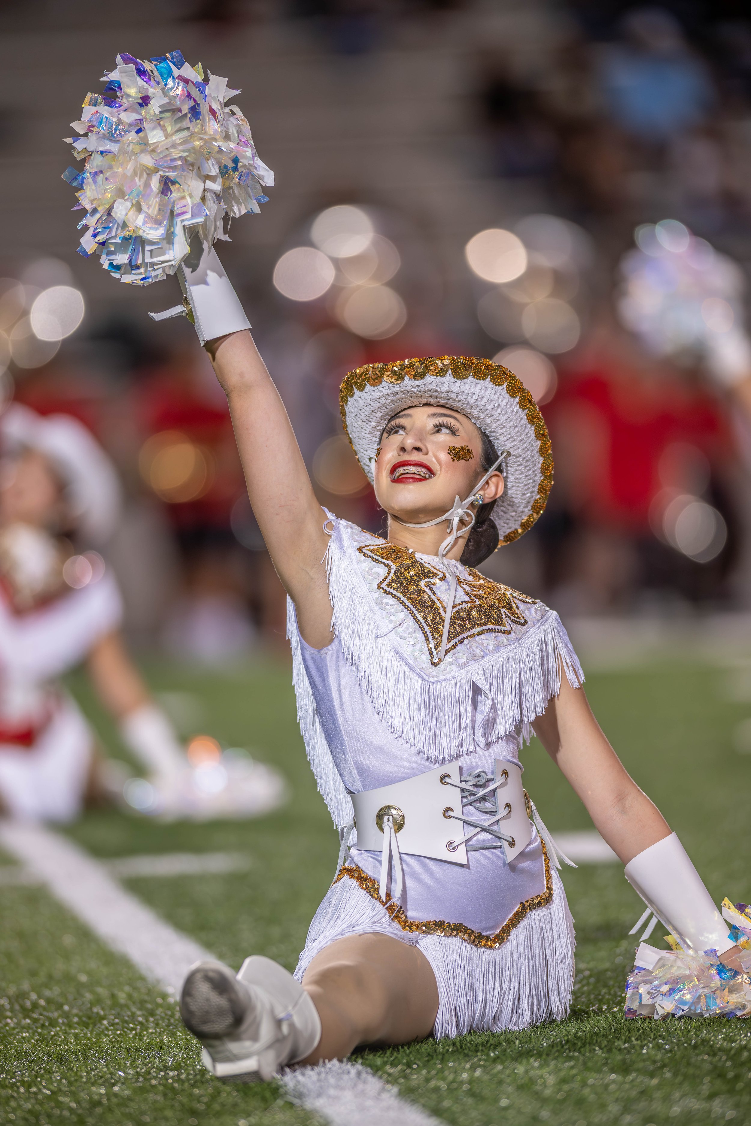 A cheerleader dressed in white with gold trim, sitting on the field during a performance, holding pom-poms, with others visible in the background.