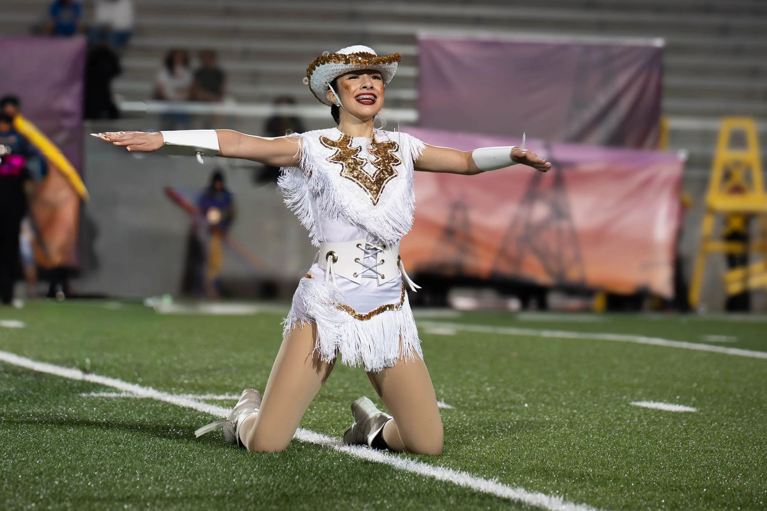 A female performer in a white fringed costume and cowboy hat kneels on a green sports field with arms outstretched, appearing emotional.