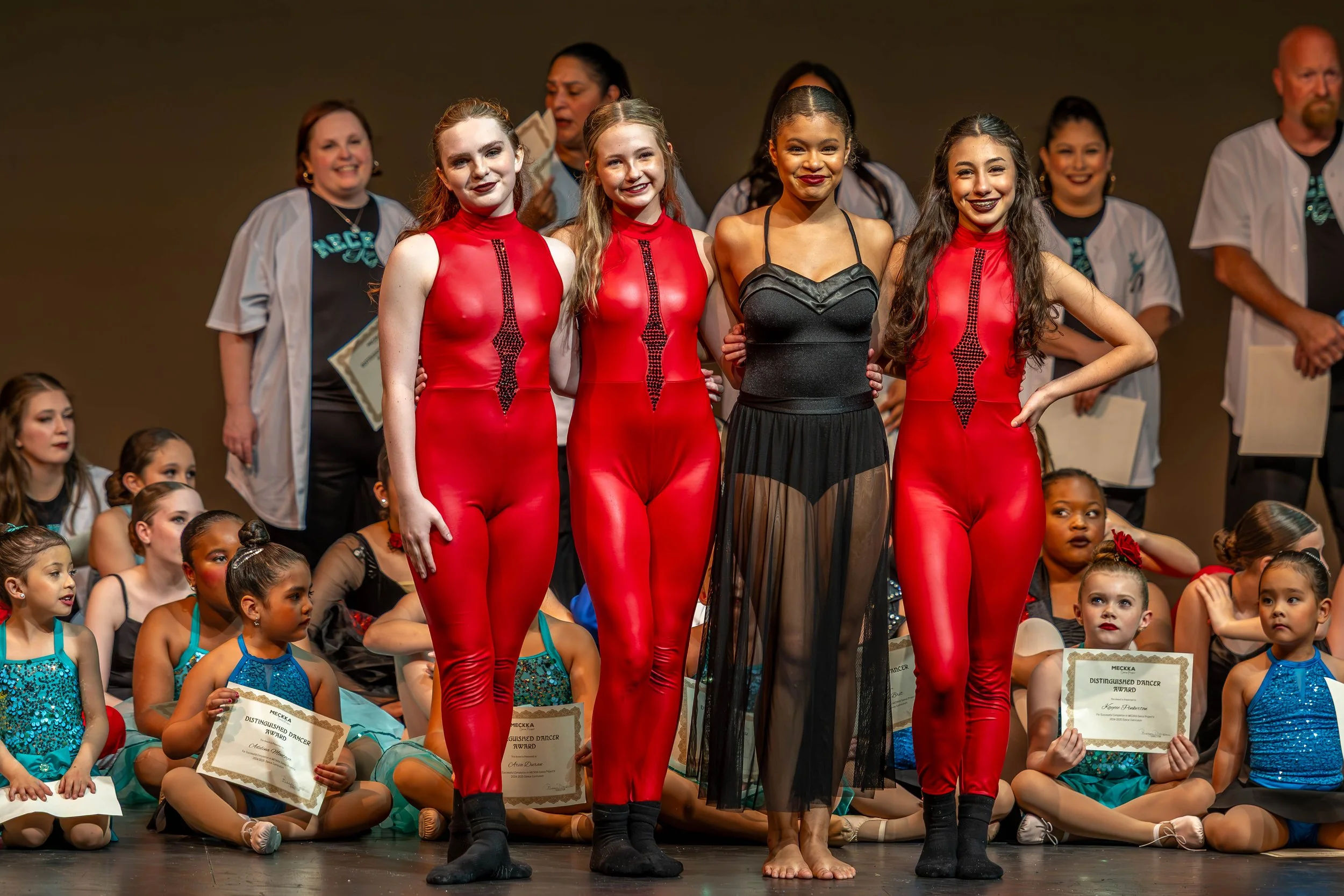 Children and young dancers gathered on stage, some holding certificates, with three teenage dancers standing in front, dressed in performance costumes, smiling.